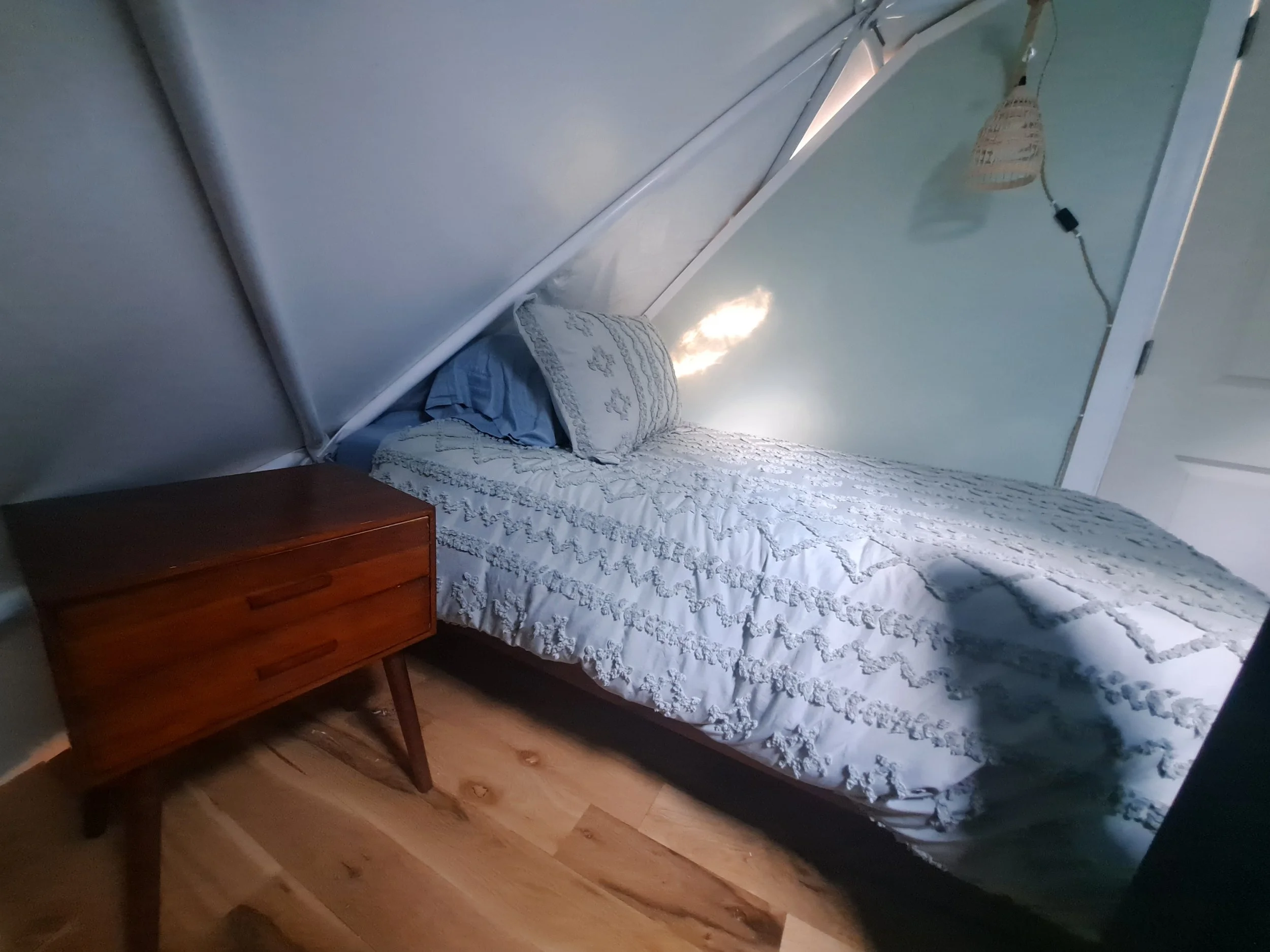 A bedroom with a bed covered in a white textured bedspread, a matching pillow and a blue pillow, a small wooden bedside table, hardwood flooring, a wall with a mirror, and a hanging woven lamp with a string light