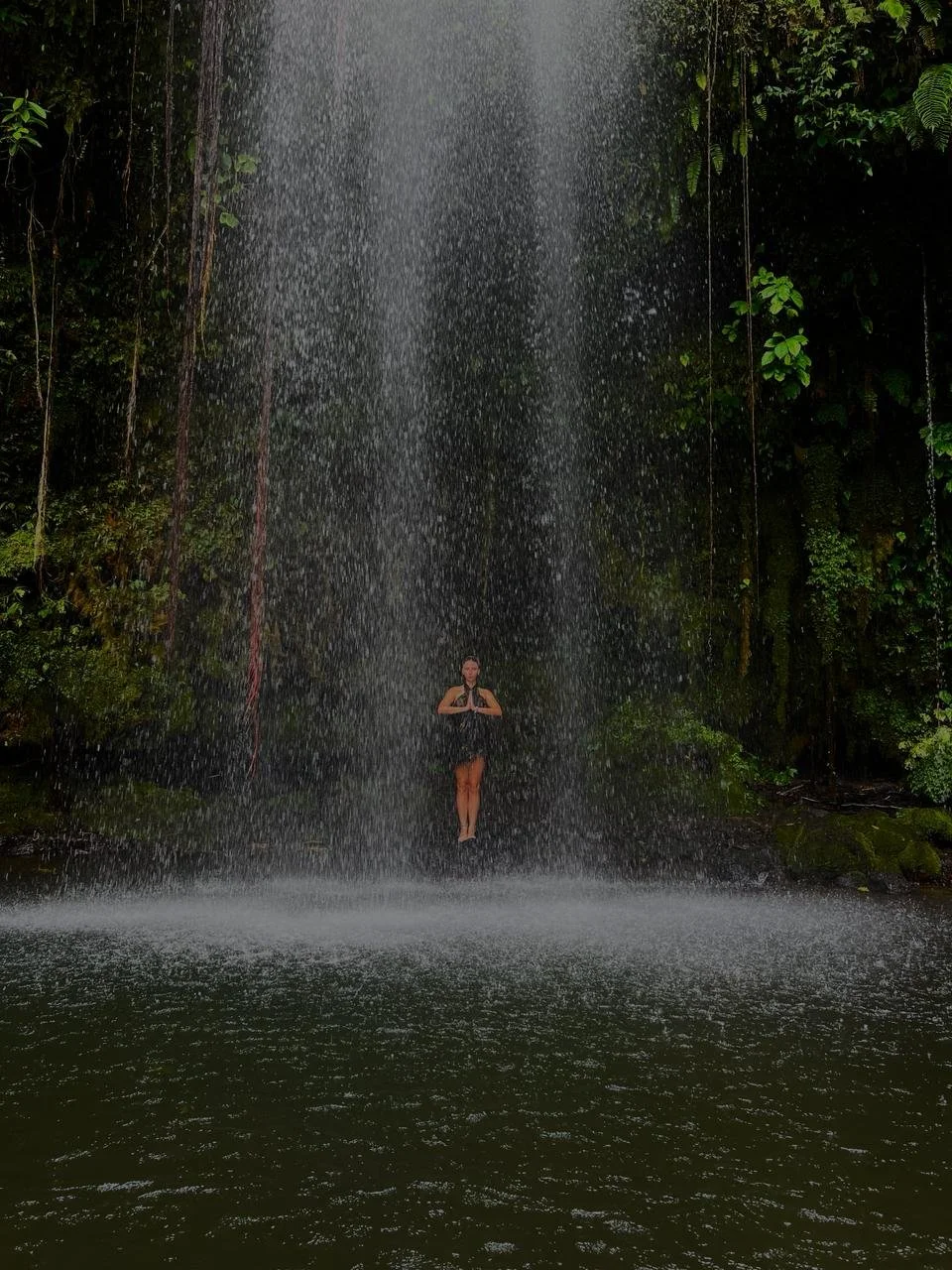 A person standing under a waterfall in a lush, green jungle, holding a prayer pose with hands pressed together.