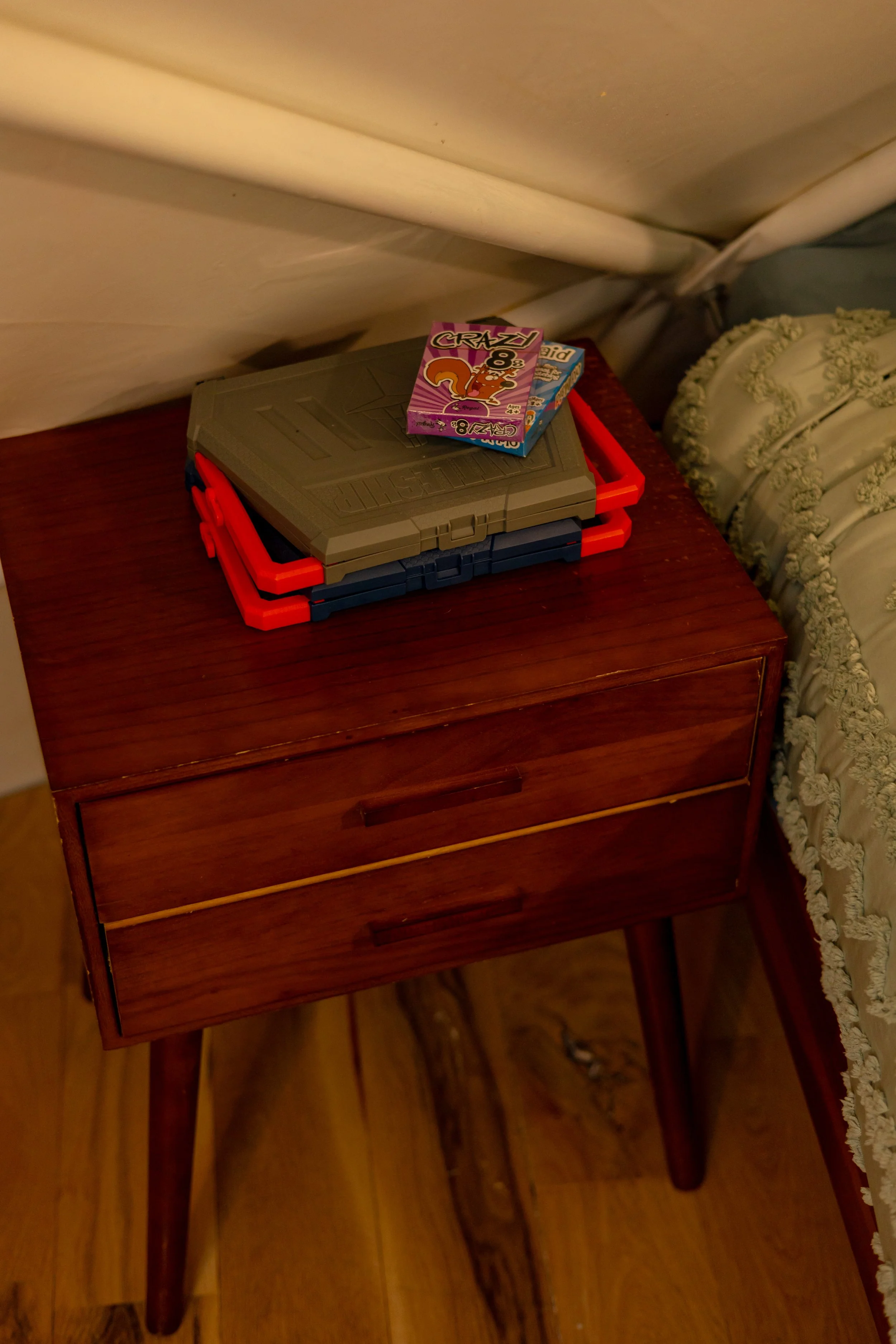 A wooden bedside table with two drawers, three game boxes on top, located next to a bed with decorative bedding.