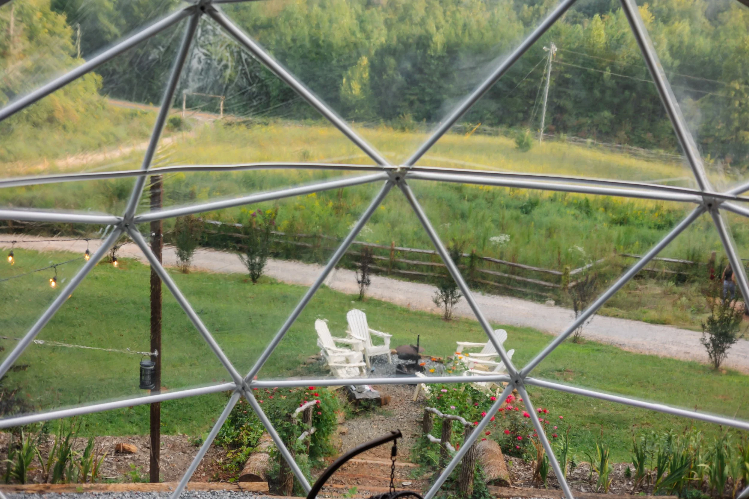 View of a backyard with a fire pit surrounded by white Adirondack chairs, a garden with colorful flowers, and a dirt pathway, seen through a geodesic dome structure.