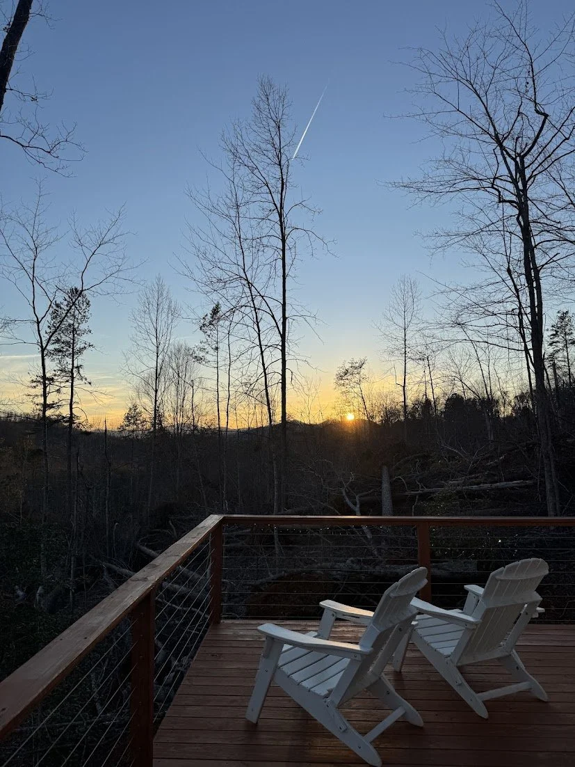 A wooden deck with two white chairs overlooking a wooded area at sunset, with bare trees and a clear sky showing a contrail.