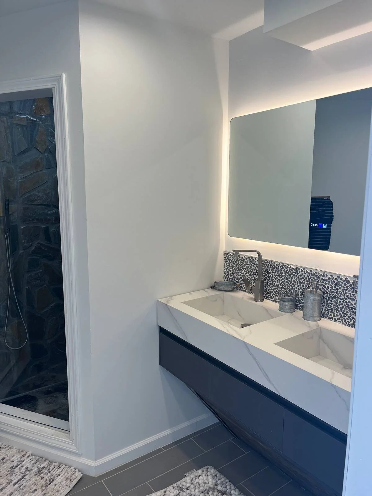 Modern bathroom with a white marble double sink vanity, a backlit mirror, and a dark blue cabinet, adjacent to a glass-enclosed shower with dark stone interior and gray floor tiles.