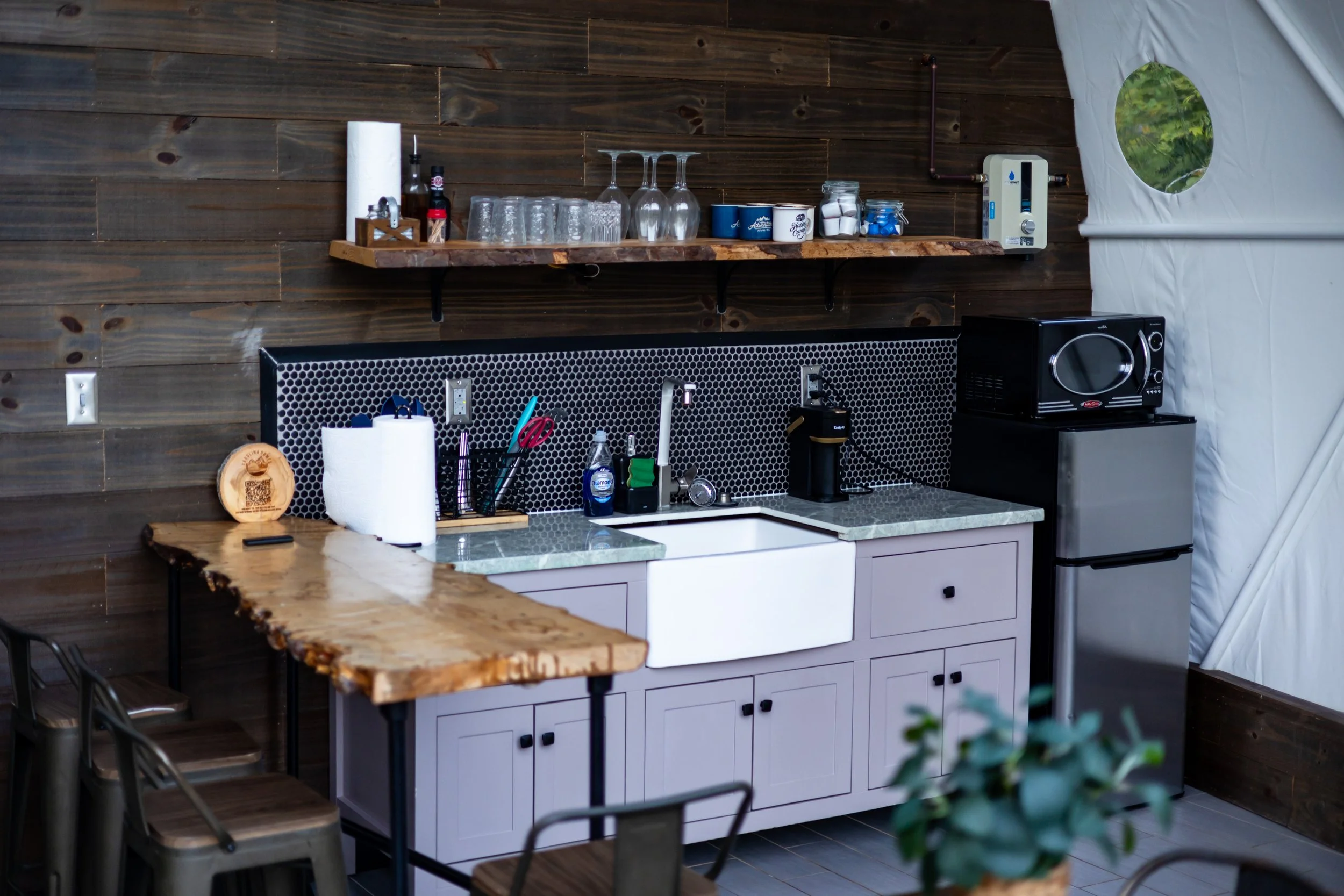 Small outdoor kitchen with a wooden countertop, light gray cabinetry, and a black honeycomb backsplash. There is a sink, faucet, paper towels, and kitchen utensil holder. On the upper wooden shelf are glasses, cups, jars, and bottles. To the right is