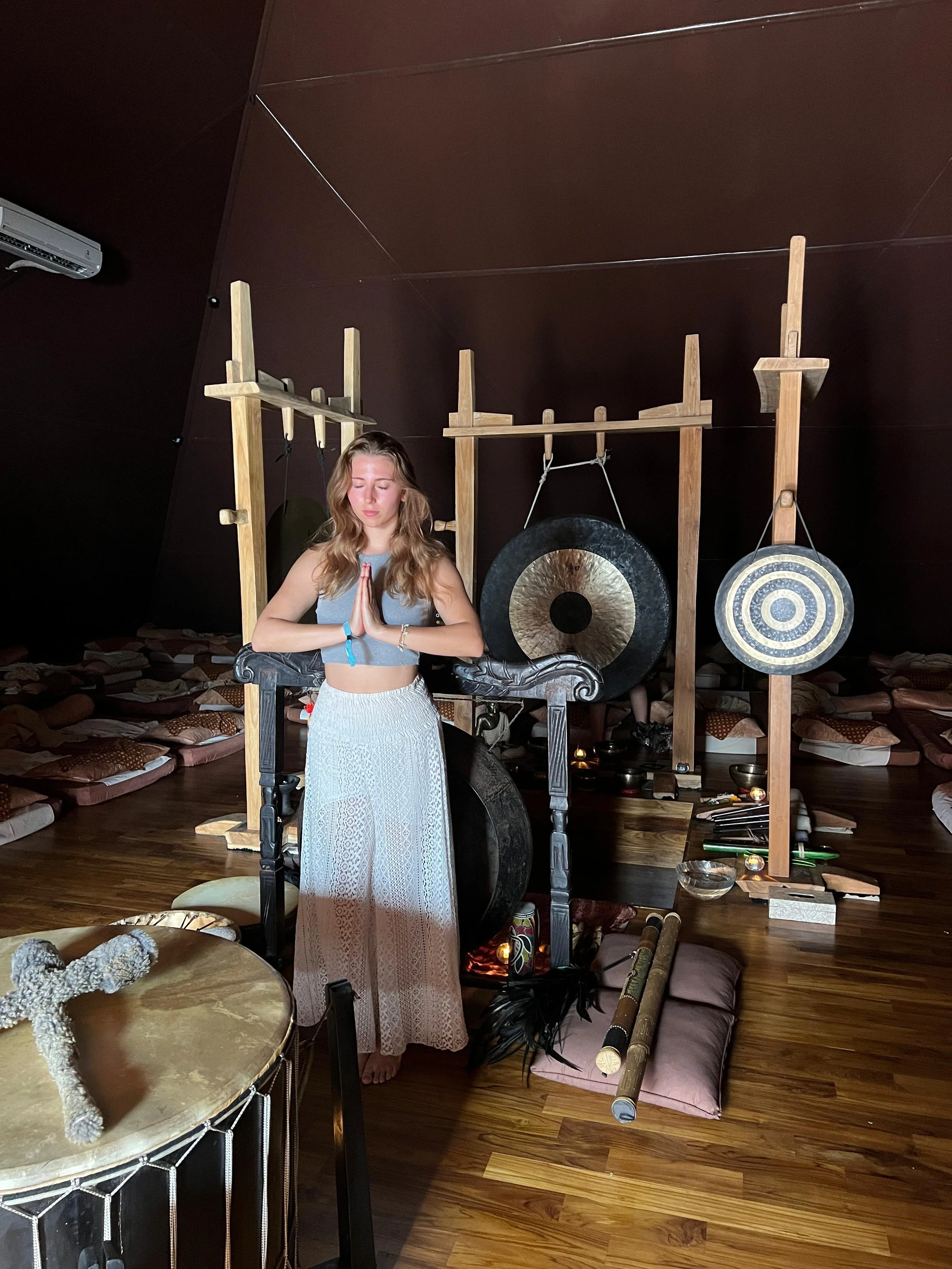 A young woman with long wavy hair standing with her hands in prayer yoga pose in front of gong instruments and meditation cushions in a dimly lit room.