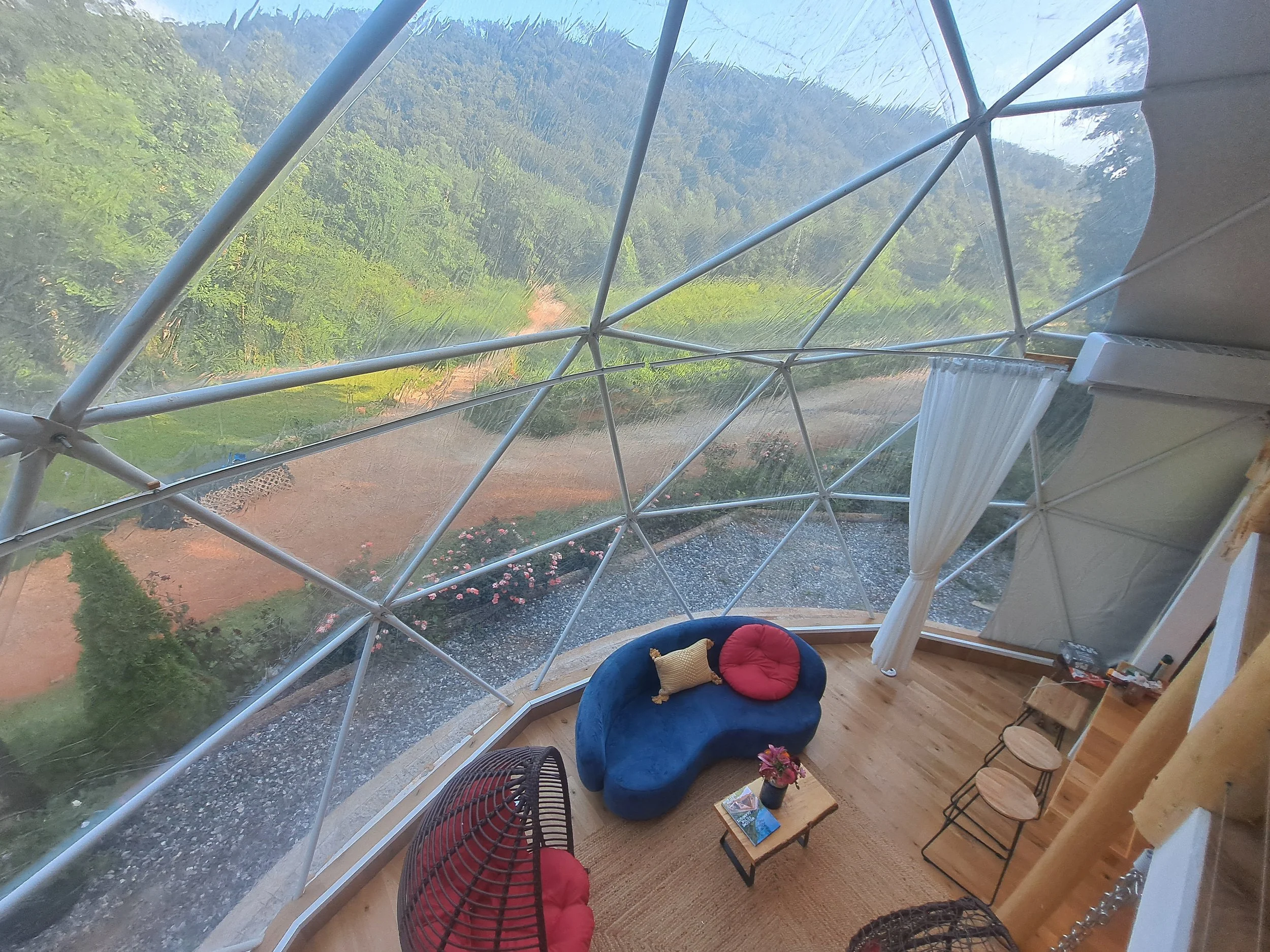 Interior of a geodesic dome with large glass windows showing a lush green landscape and mountains outside. Inside, there is a curved blue sofa with a red round cushion and a small pillow, a wooden coffee table with books and flowers, and a white curt