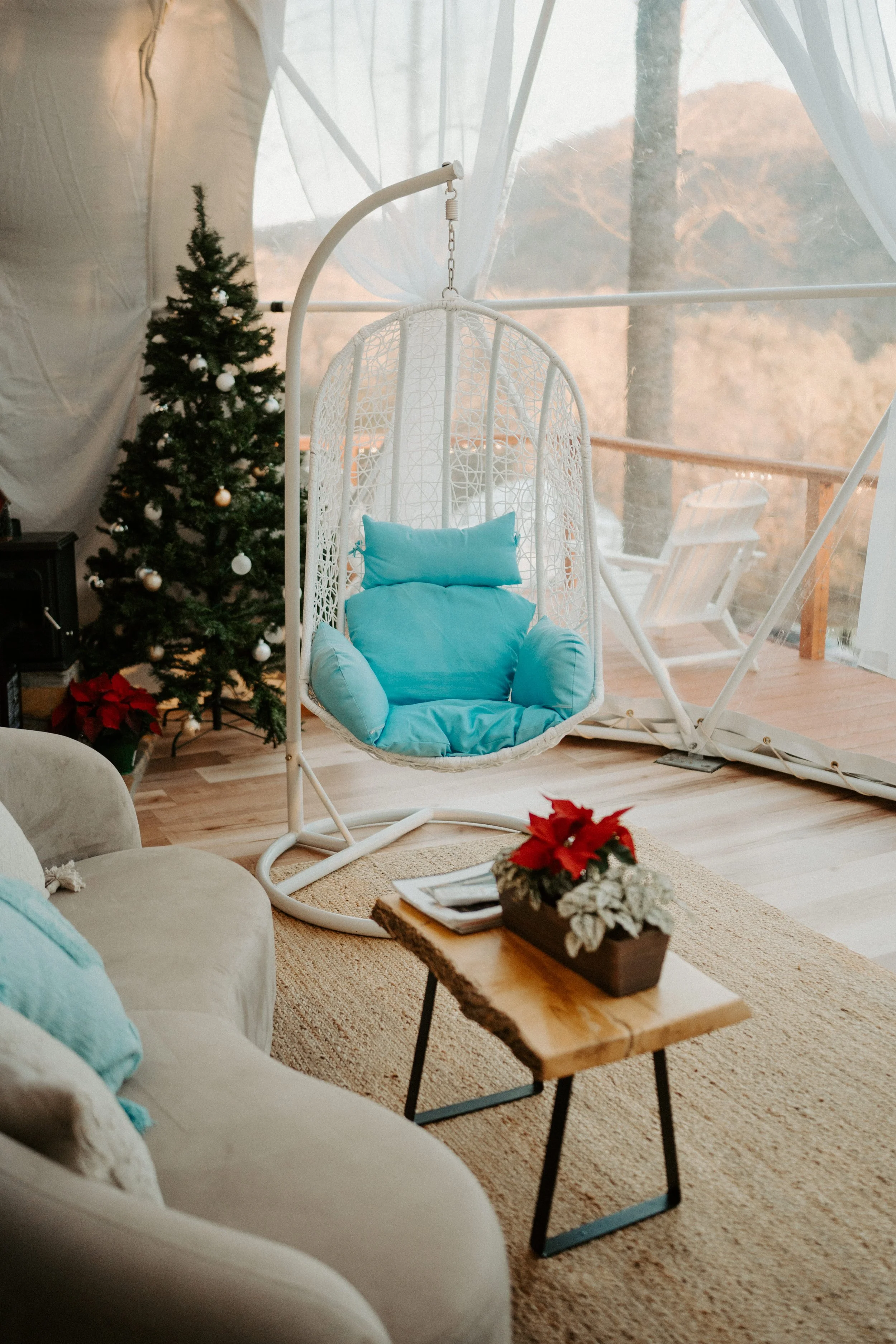 A cozy indoor space with a beige sofa, light blue cushions, a wooden coffee table with poinsettia plant, a white hanging egg chair with blue cushions, a decorated Christmas tree, and a scenic view of trees and mountains through a porch or window.