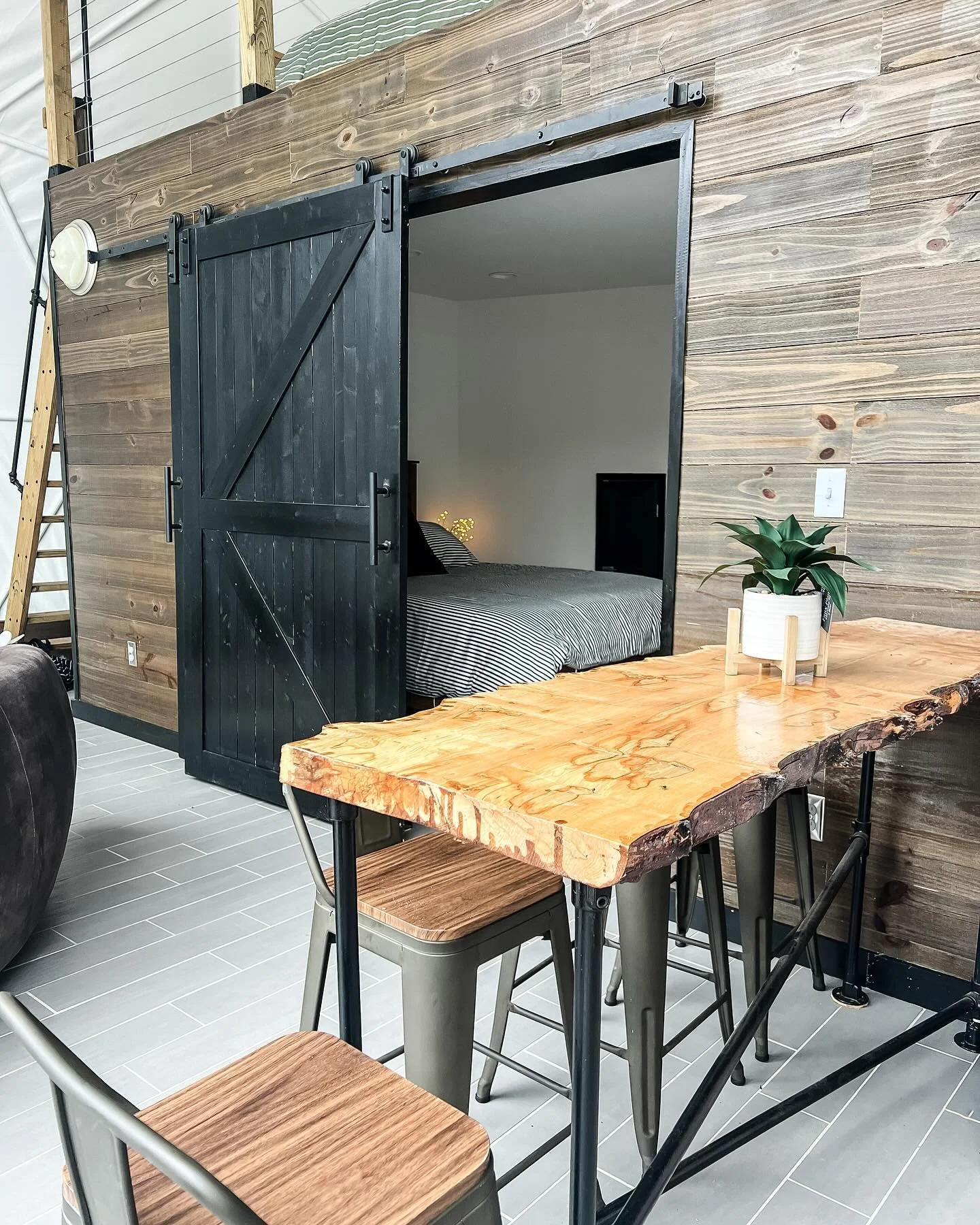 A room with a rustic wooden table and metal chairs, a black sliding barn door leading to a bedroom with a bed and striped bedding, a potted plant on the table, and wood-paneled walls.