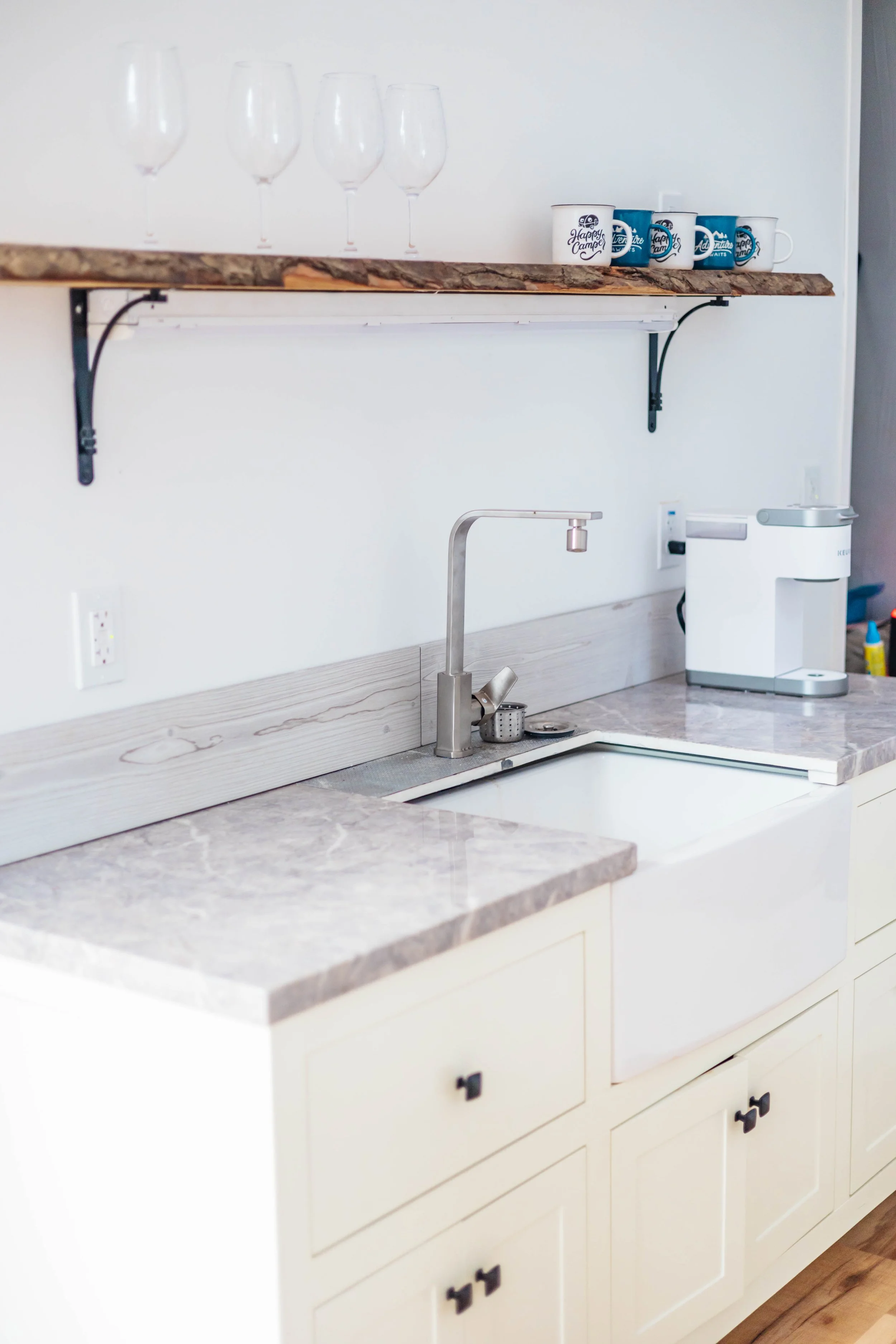 Clean kitchen with a marble countertop, white cabinetry, a modern stainless steel sink and faucet, and a coffee machine on the right. Glasses and mugs are on a shelf above the sink.