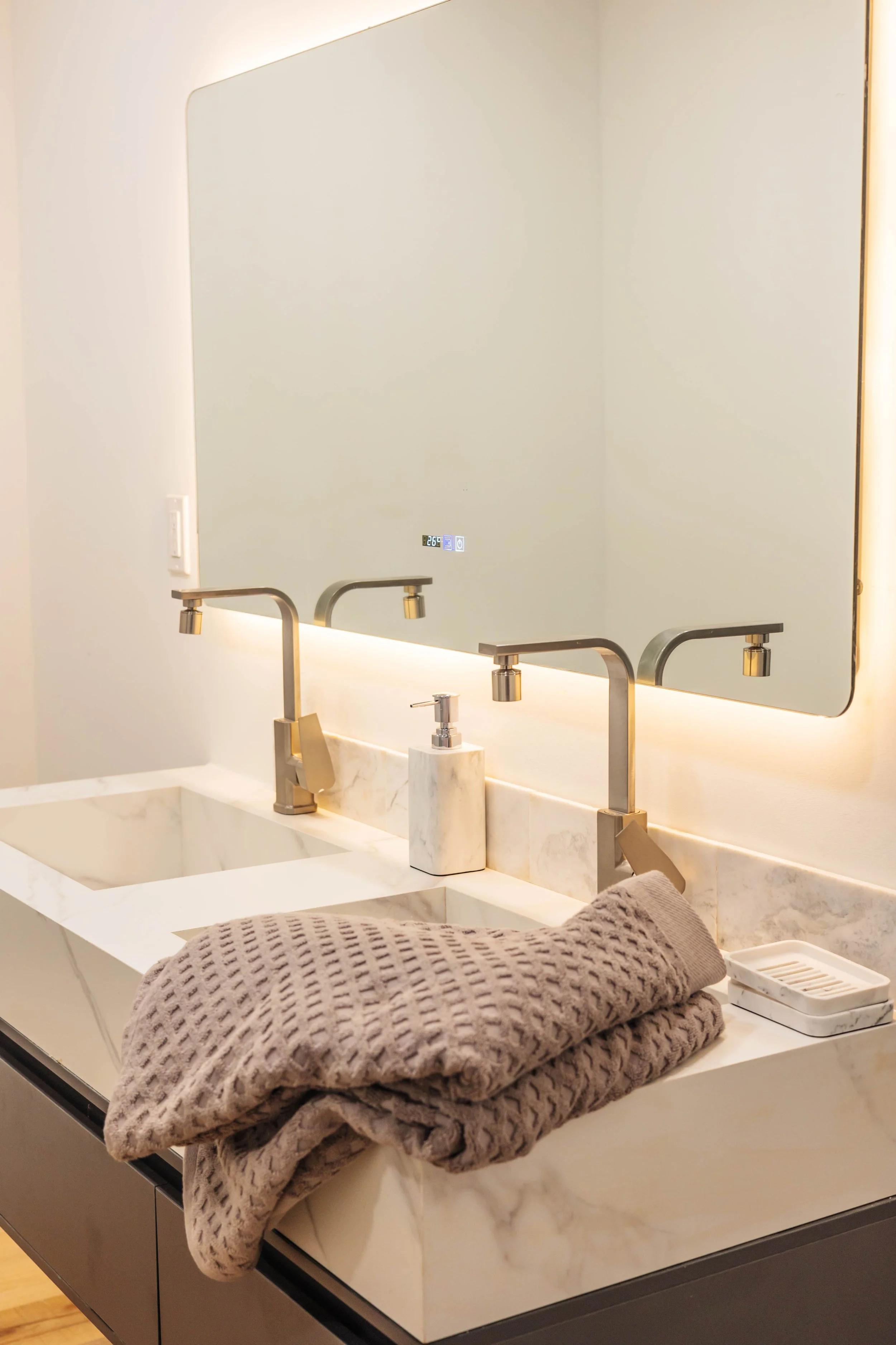 Bathroom double sink with marble countertop, two modern faucets, a soap dispenser, a bath towel, and a large illuminated mirror.