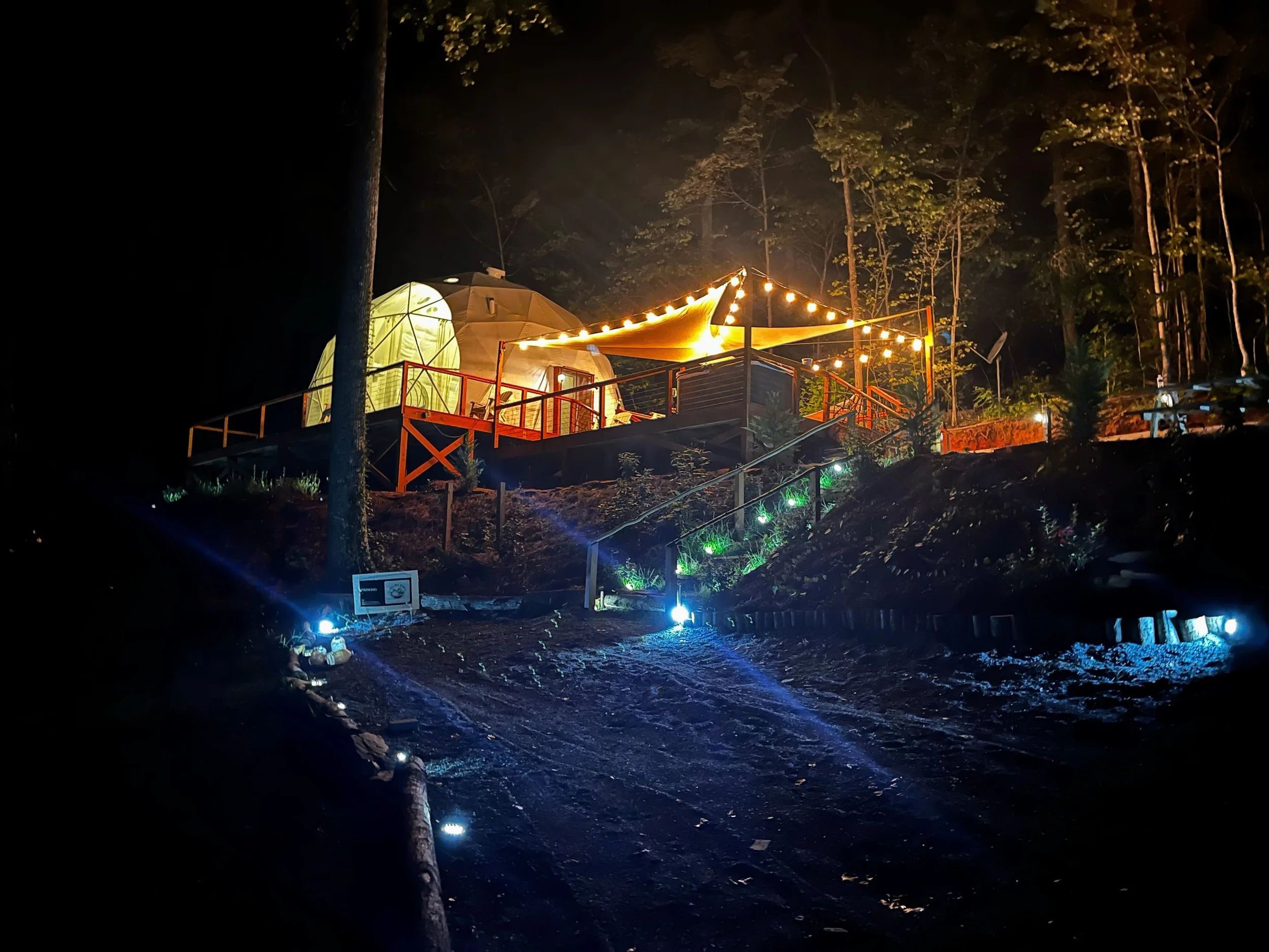 A campsite with a yellow dome tent, decorated with string lights, on a raised wooden platform in a forest at night, illuminated by colorful outdoor lighting.