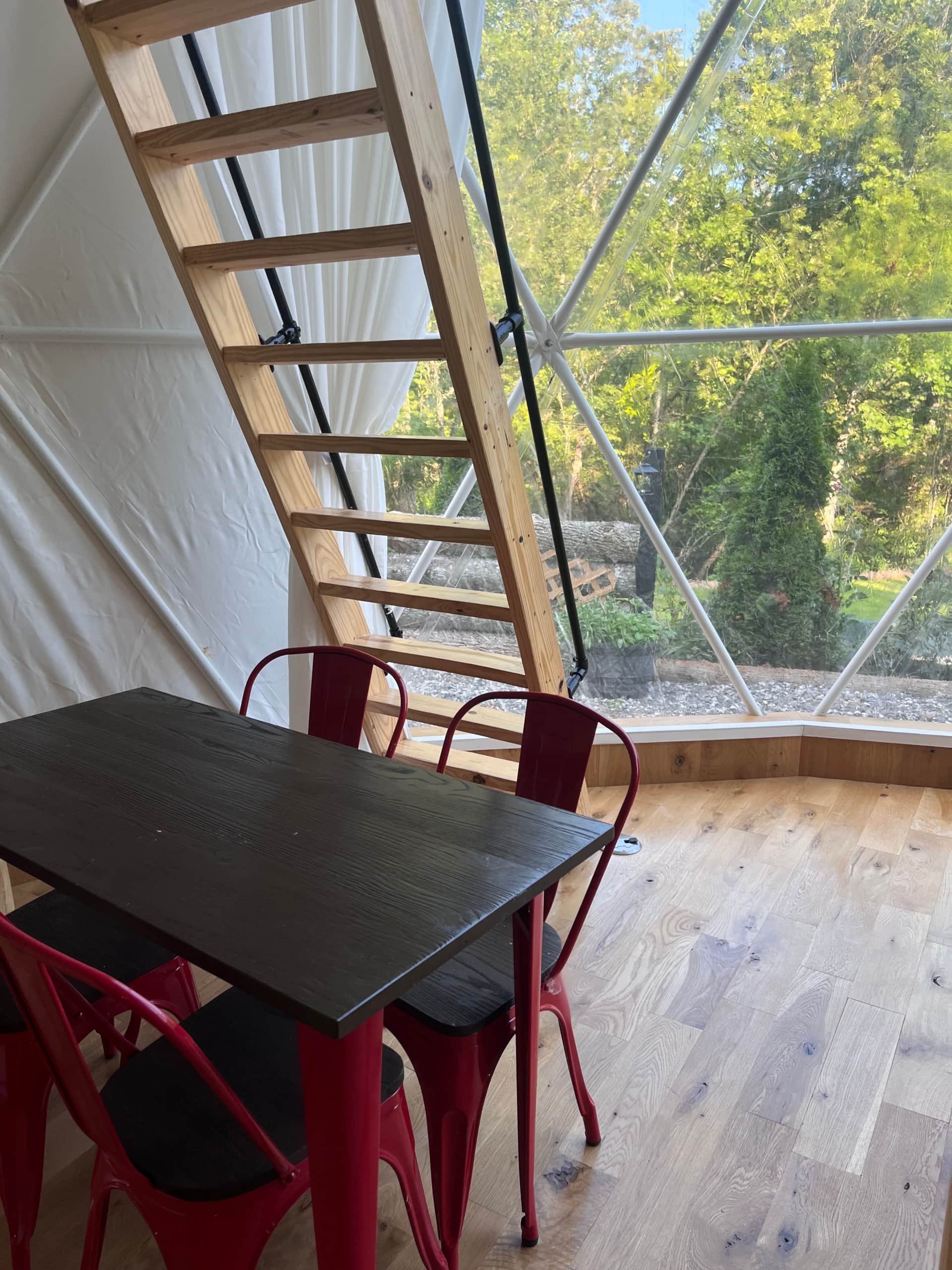 Interior of a wooden dome structure with a black table and four red chairs, a wooden staircase, and a large window showing green trees outside.