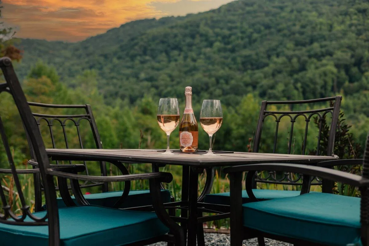 A table with a bottle of rosé wine and two glasses filled with rosé, set outdoors with a scenic view of green hills and a sunset or dawn sky in the background.