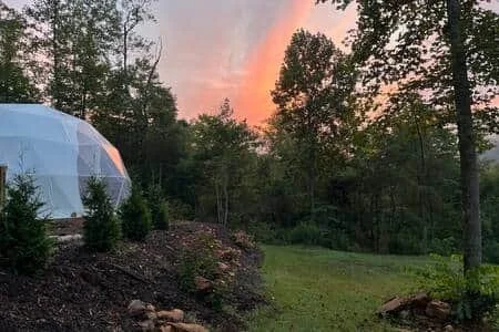 Sunset over a wooded landscape with a geodesic dome on the left and grassy area in the foreground.