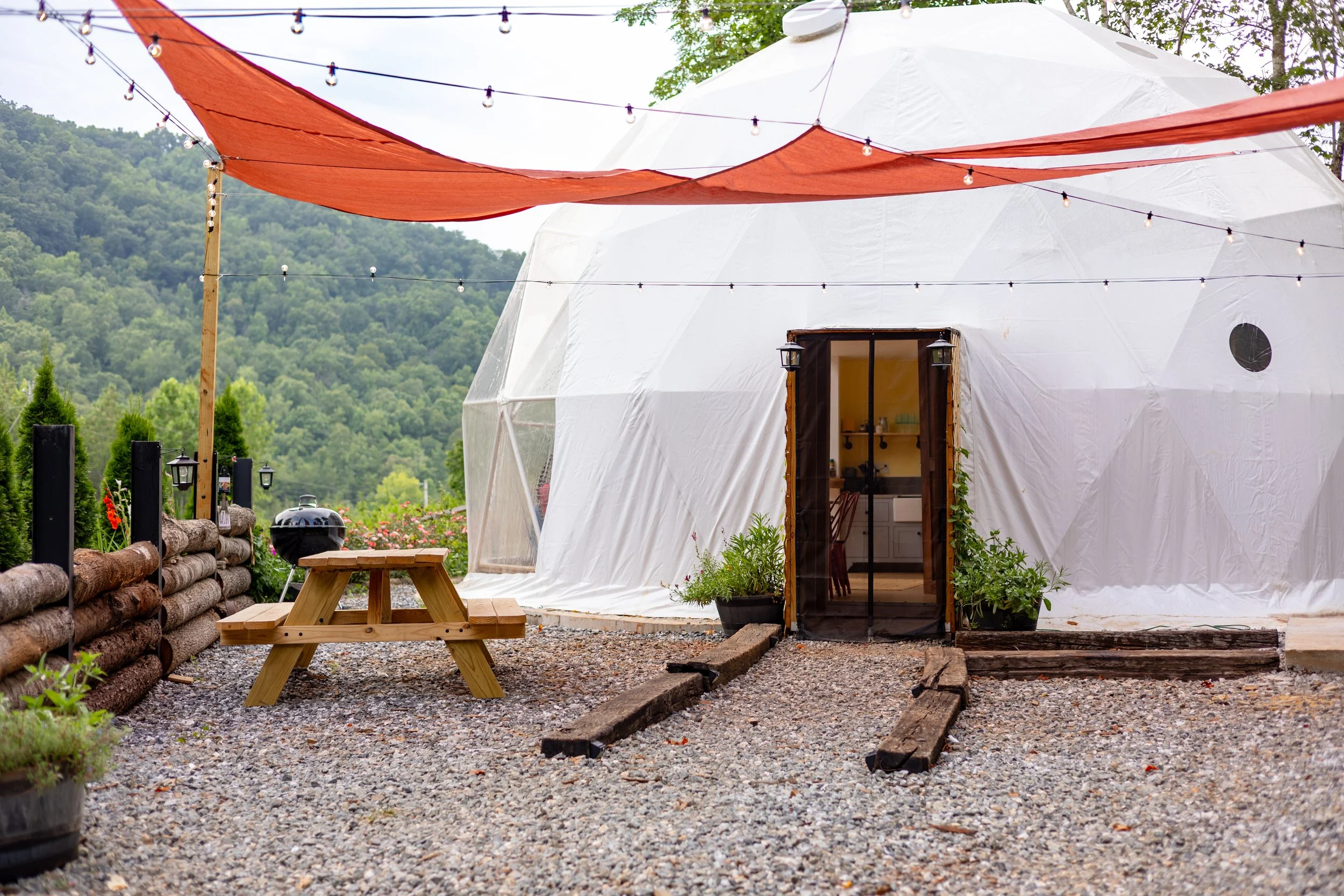 Outdoor scene with a geodesic dome tent, string lights, a picnic table, potted plants, and a background of green hills.
