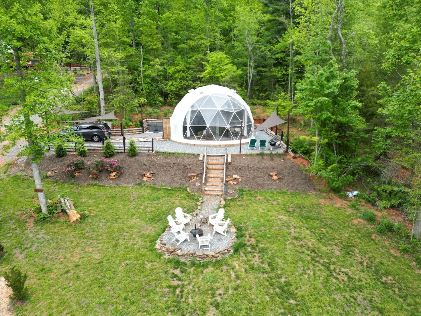 A backyard with a green lawn, a fire pit surrounded by white Adirondack chairs, a pathway leading to a white geodesic dome, and surrounded by trees.
