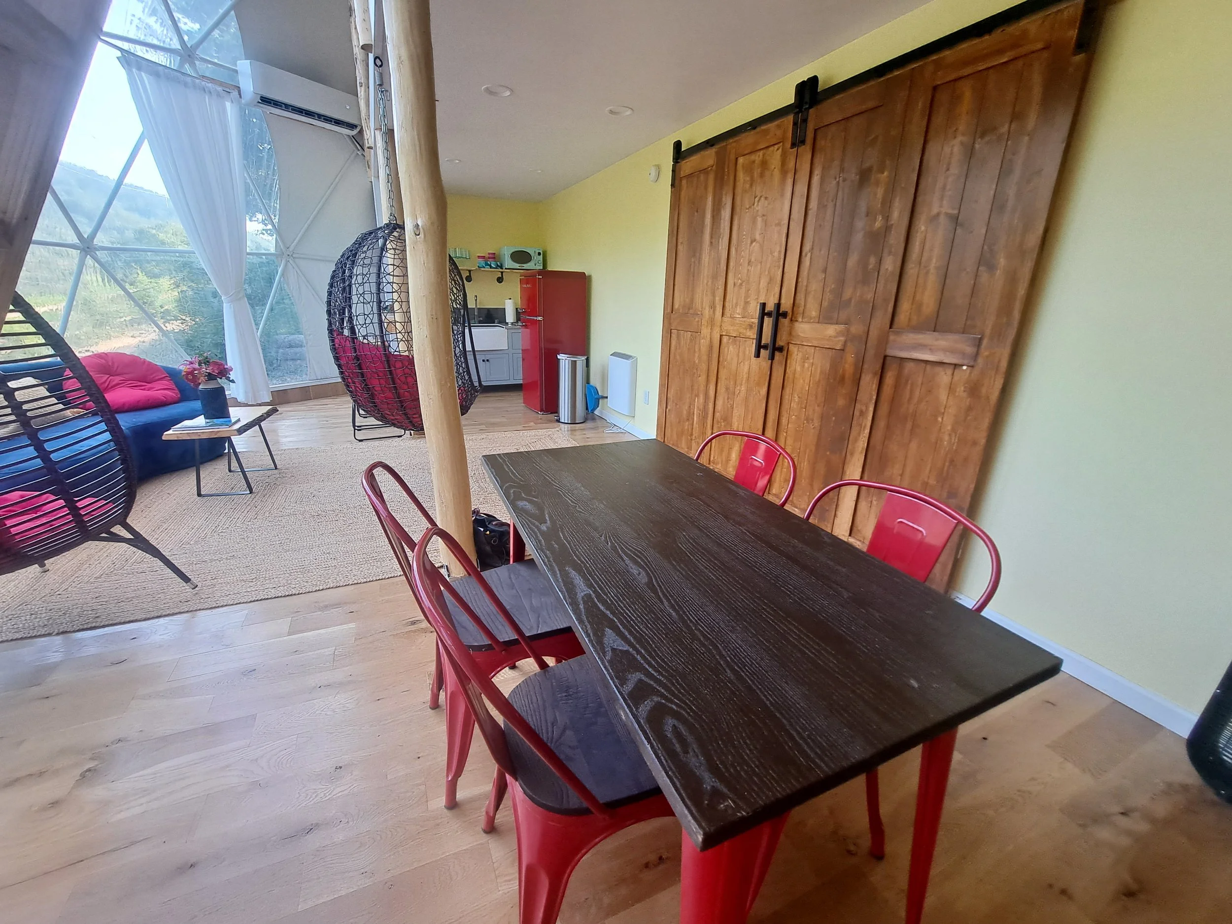 A dining area with a dark wooden table and red chairs, a large wooden sliding door, a small kitchen with a refrigerator and microwave, a cozy sitting area with a navy blue couch with pink pillows, hanging chairs, and a large window with white curtain