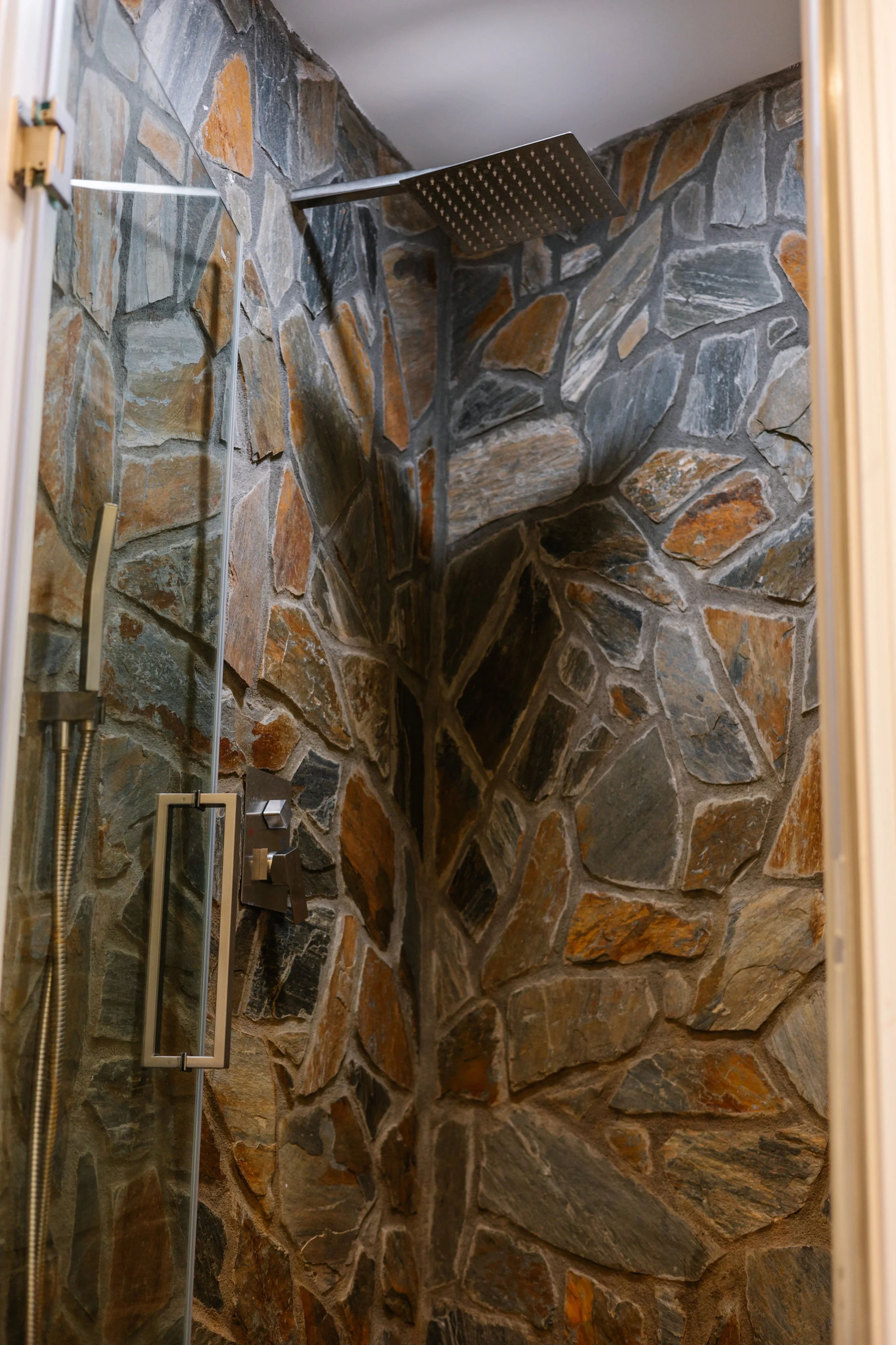 A walk-in shower with stone tile walls, a large square showerhead mounted on the ceiling, and a glass door with a metal handle.