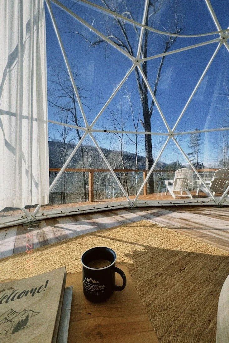 Inside a geodesic dome with large windows showing a snowy outdoor view, including trees and a deck with Adirondack chairs. On a wooden table, there's a black mug filled with coffee and a welcome brochure.