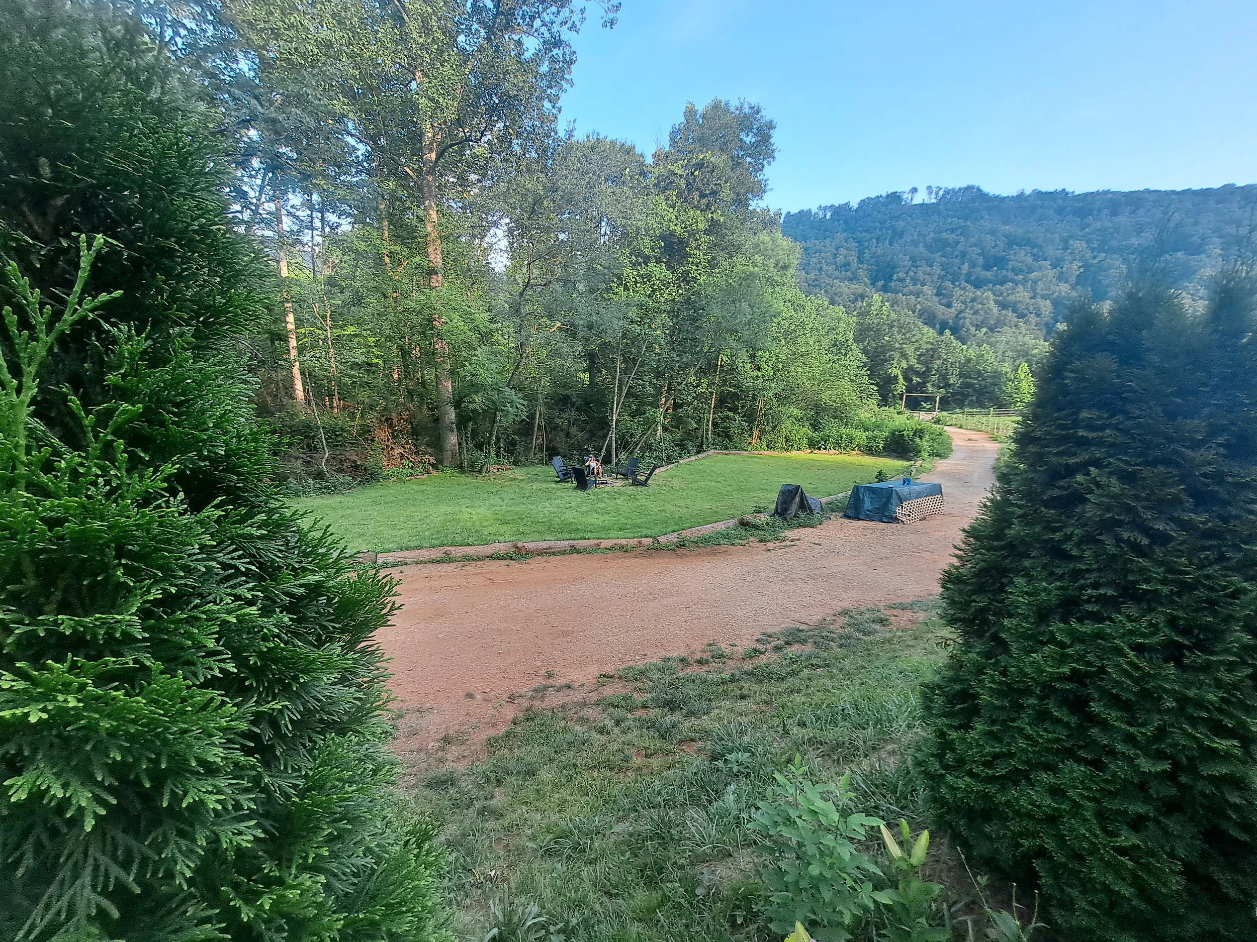 A serene outdoor scene with a dirt pathway, green grass, trees, and hills in the background. There are outdoor chairs and tables, some covered with tarps, under a clear blue sky.