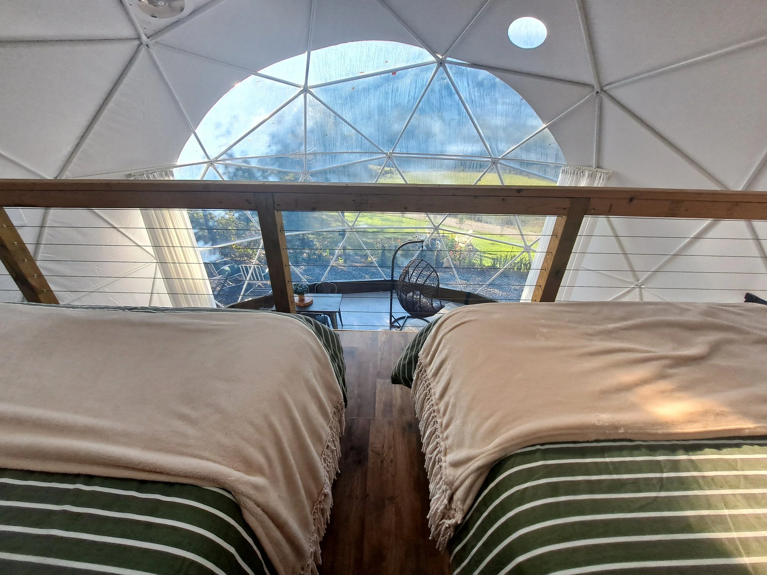 Inside a geodesic dome with two beds covered in striped bedding and beige blankets, overlooking scenic outdoor landscape through large semi-circular window.