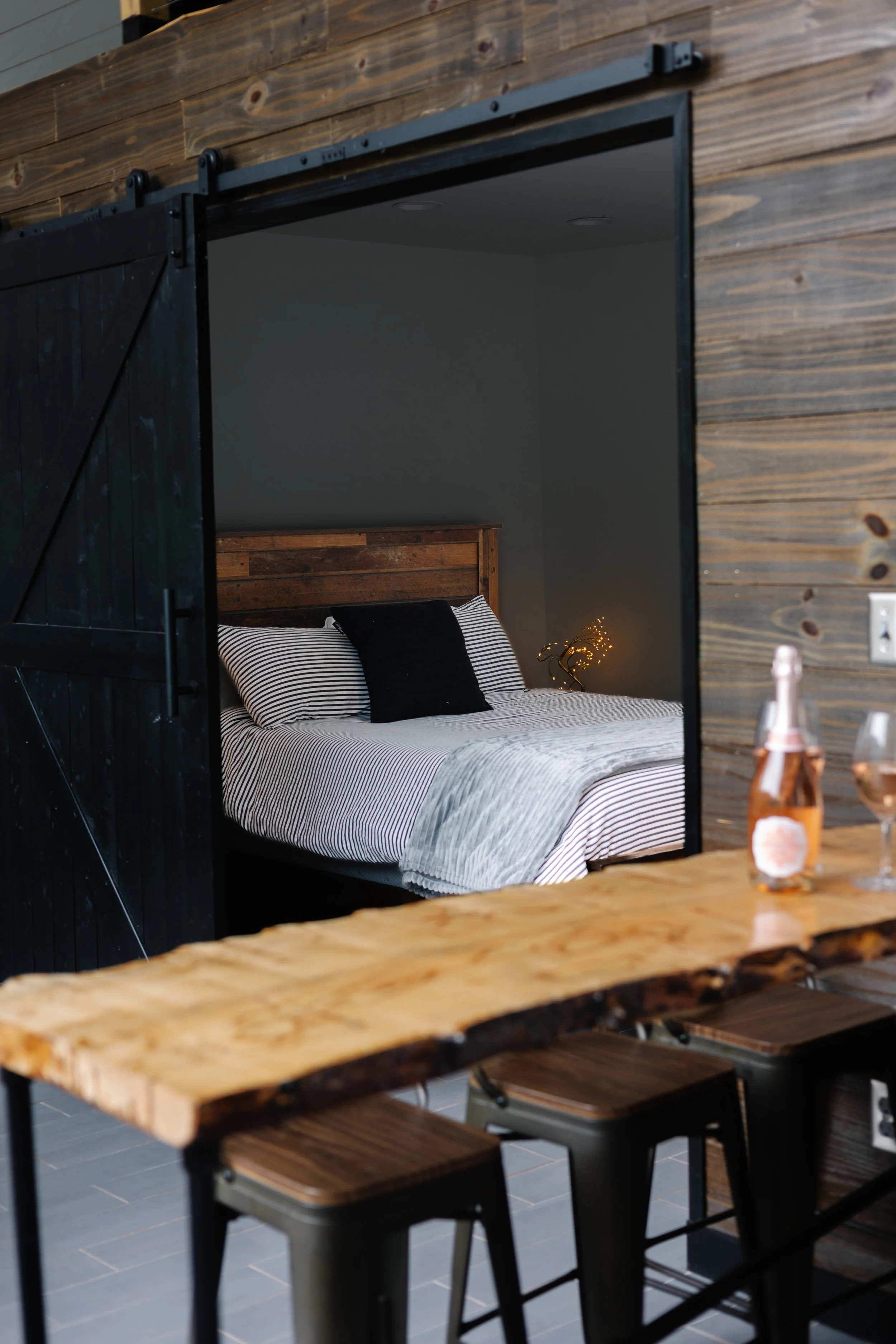 View of an interior bedroom through a black barn door, featuring a bed with striped bedding and pillows, and a bedside lamp with string lights. In the foreground, a rustic wooden table with a bottle and glass of wine.