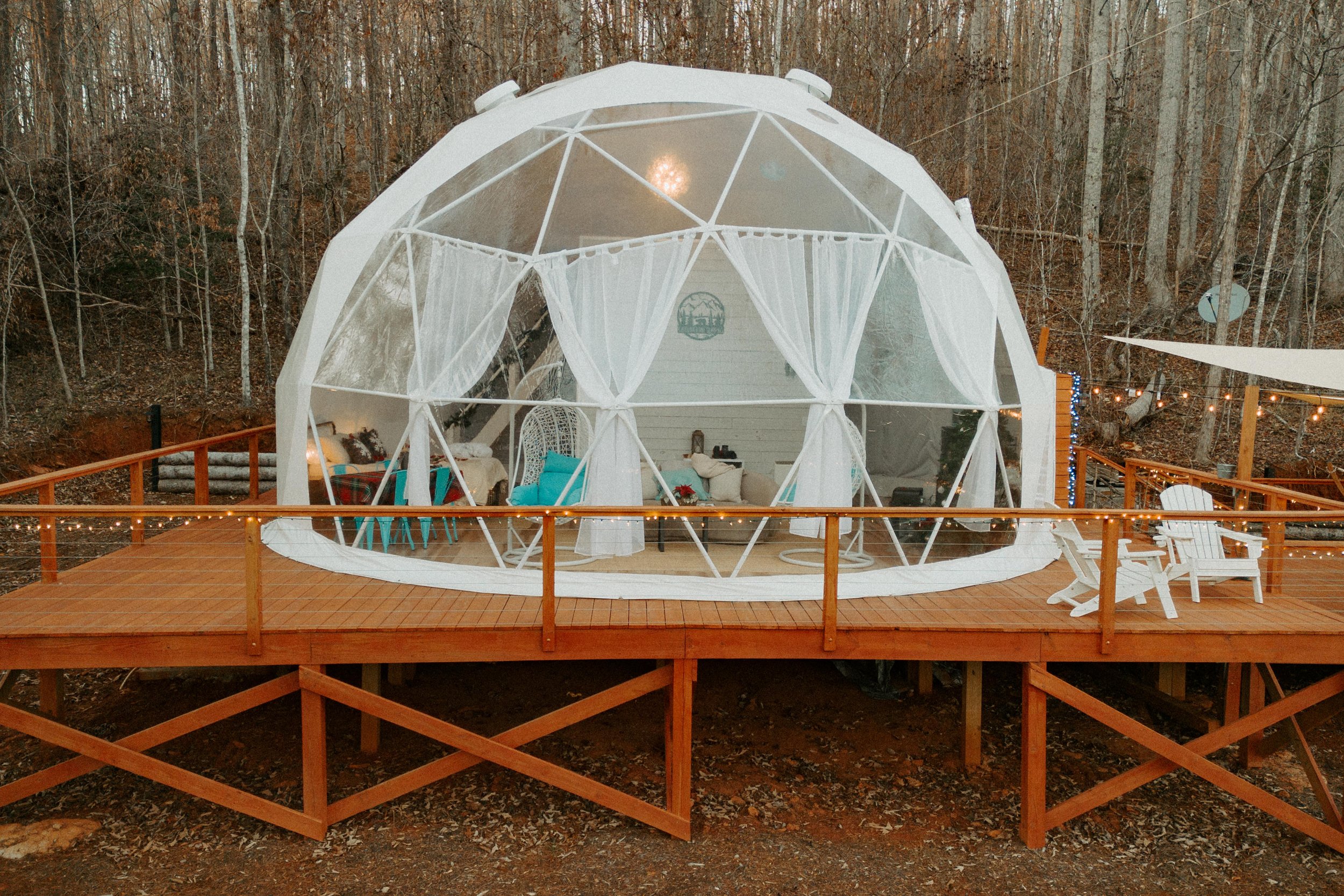 A white geodesic dome on a wooden deck with outdoor furniture, string lights, and curtains, set in a wooded area.