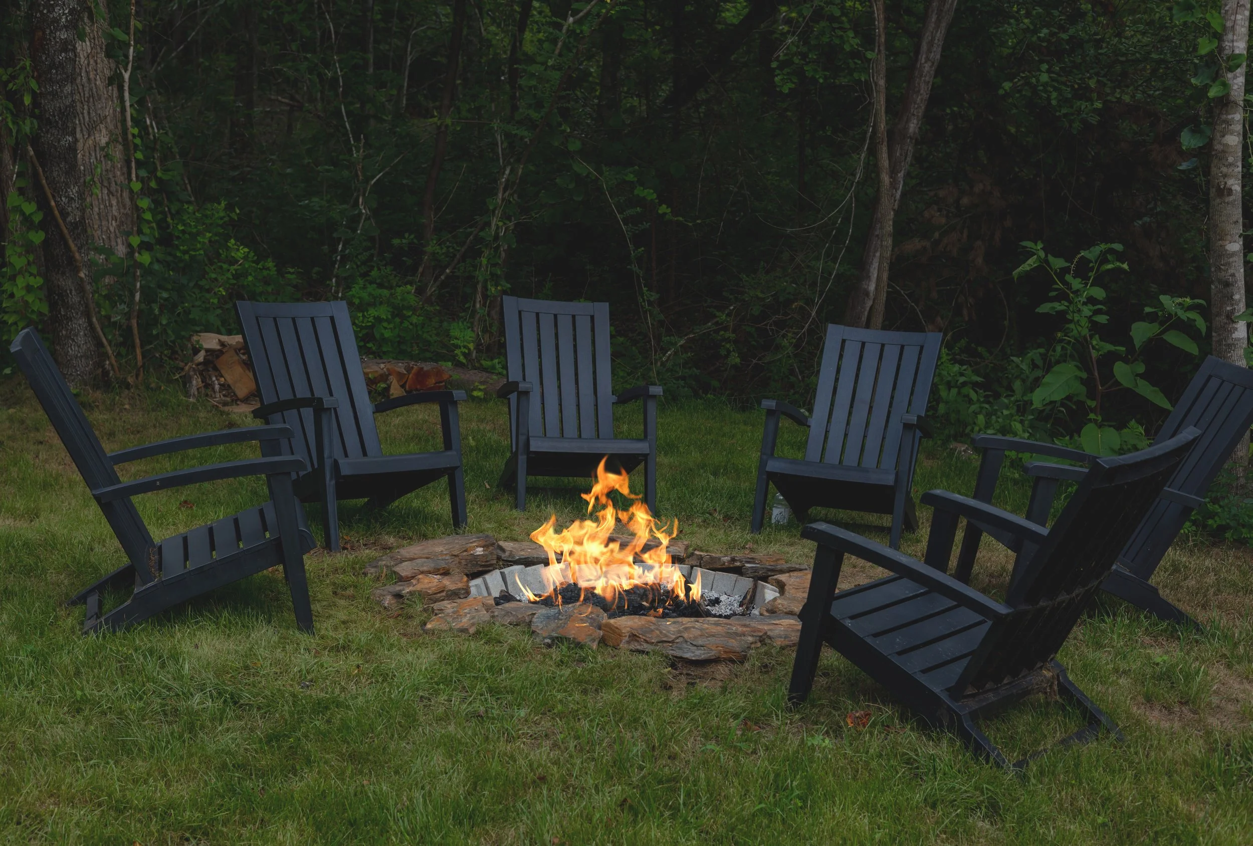 Six black Adirondack chairs arranged in a circle around a fire pit on a grassy area, with trees and dense green foliage in the background.