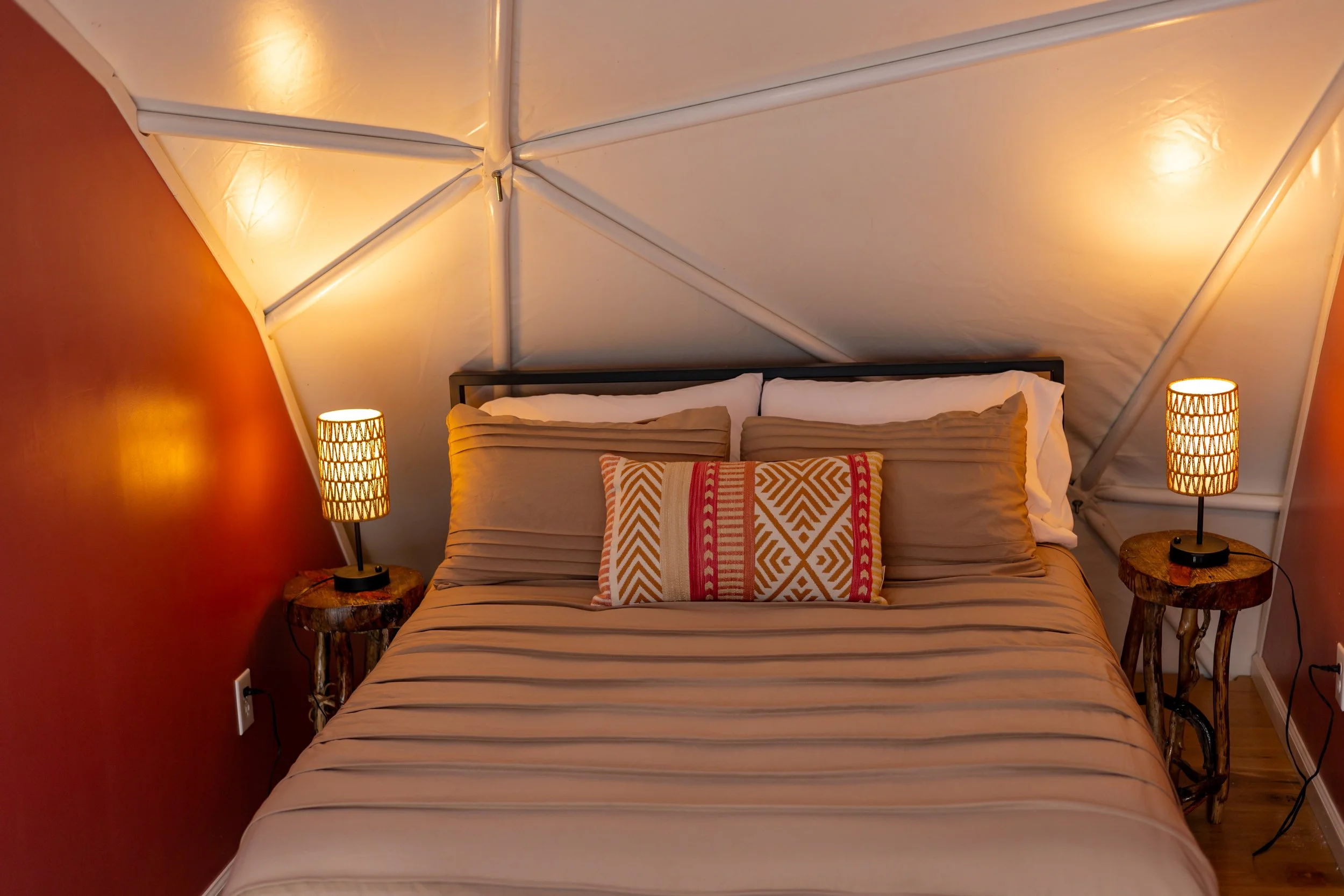A cozy bedroom with a bed, beige striped bedding, and decorative pillows, flanked by two rustic wooden nightstands with matching lamps, inside an A-frame structure with white walls and warm lighting.
