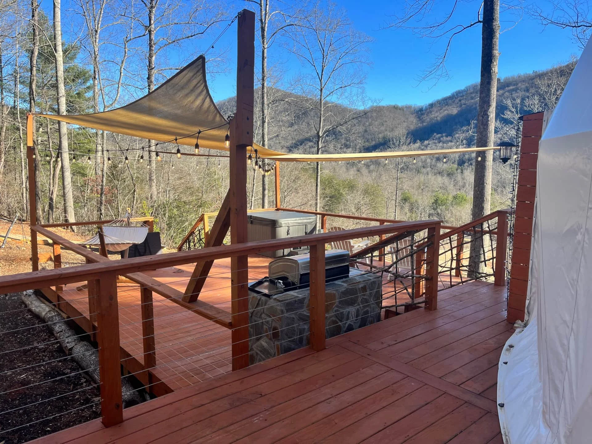 Wooden deck with string lights, a container, and a small grill against a backdrop of trees and mountains on a clear day.