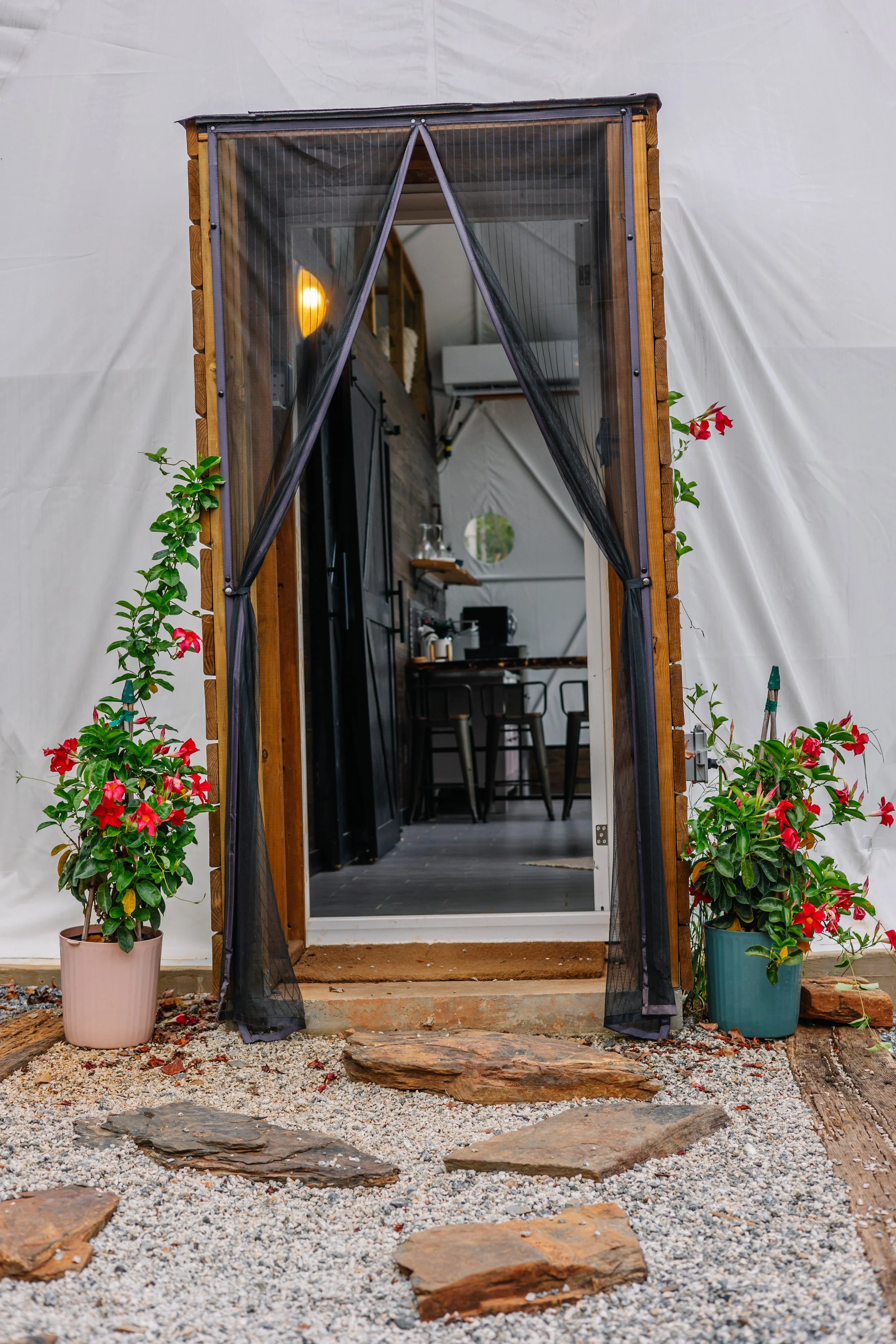 Entrance to a tiny house with black curtains, potted pink and red flowers on both sides, and a stone path leading inside.