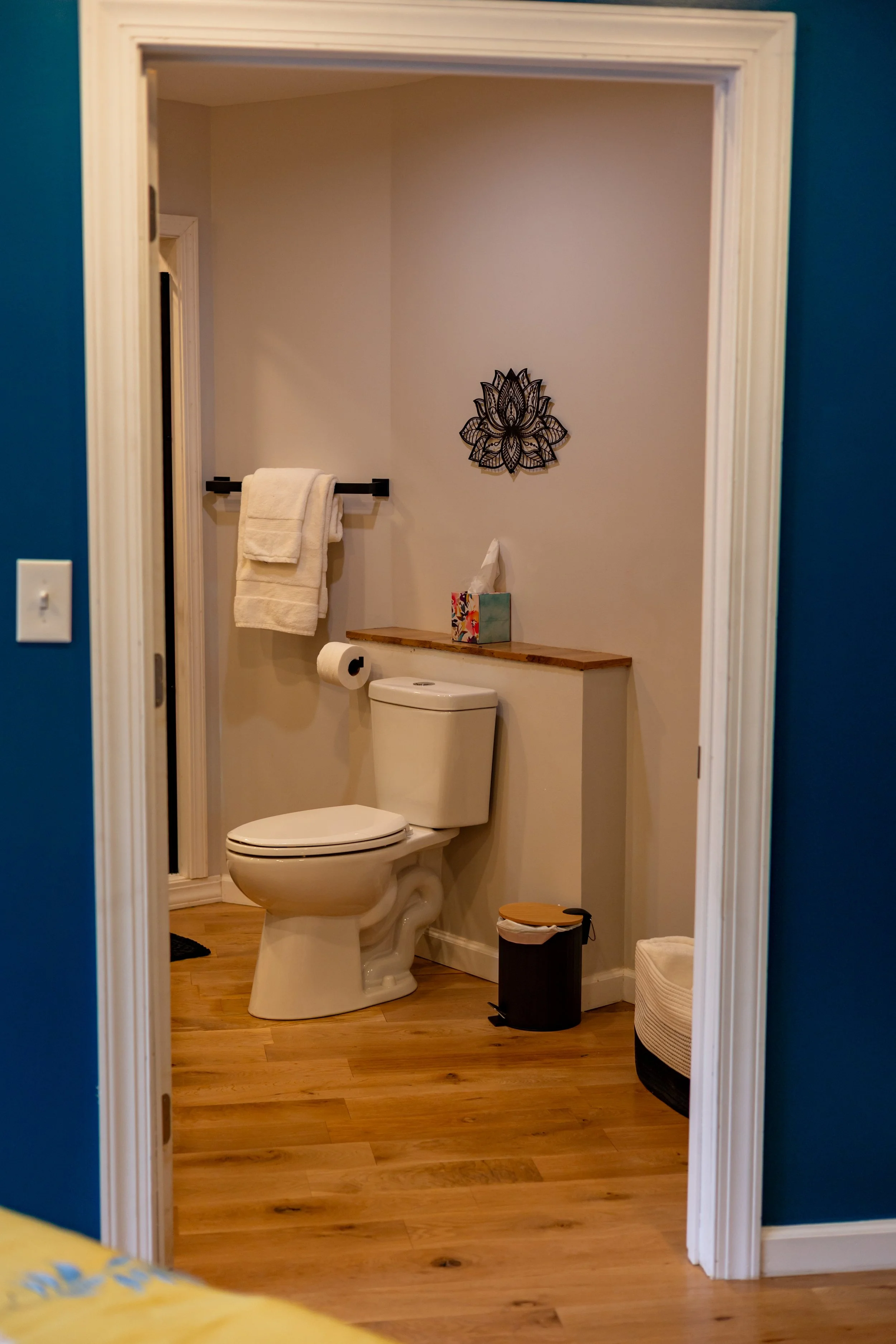 The image depicts a small bathroom with a white toilet, a wooden shelf holding a box of tissues, a roll of toilet paper, and decorative wall art. A black towel rack with white towels is on the wall, and the floor is wooden. The bathroom is viewed thr