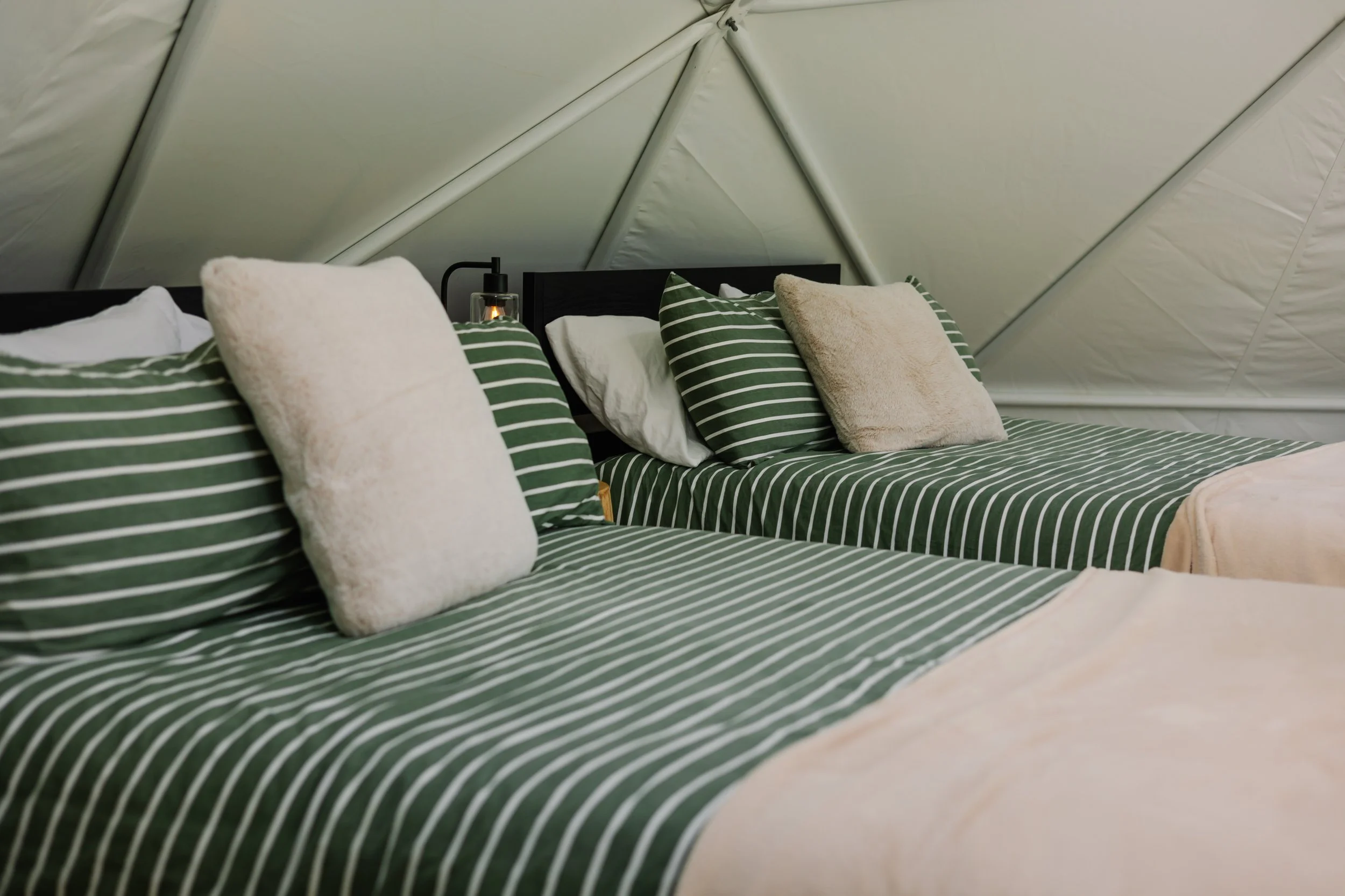 Two beds with green and white striped bedding, beige and white pillows, inside a tent with cream-colored walls.