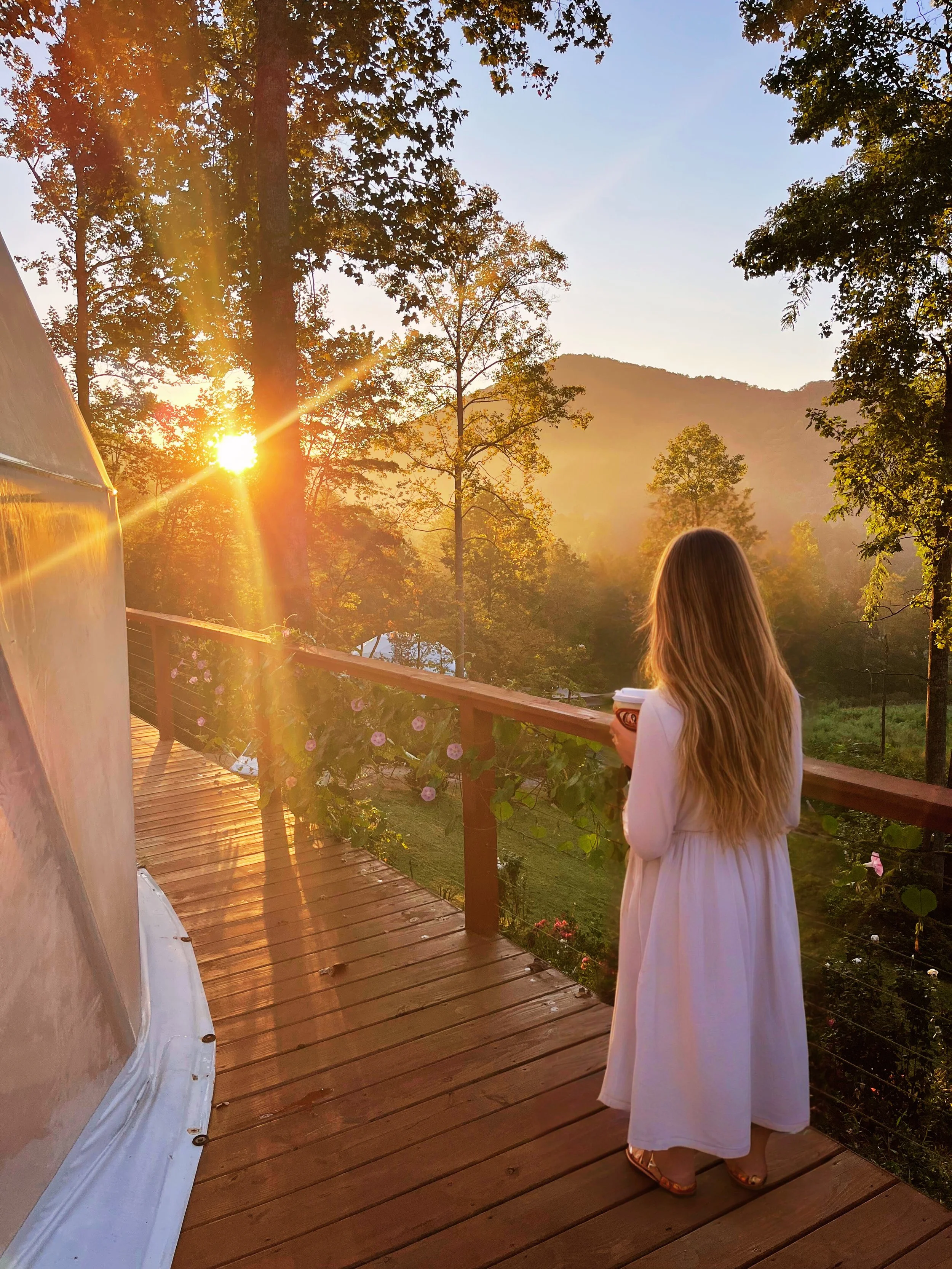 A woman with long blonde hair in a white dress standing on a wooden balcony, holding a cup, looking at the sunrise over a forested landscape with hills in the background.