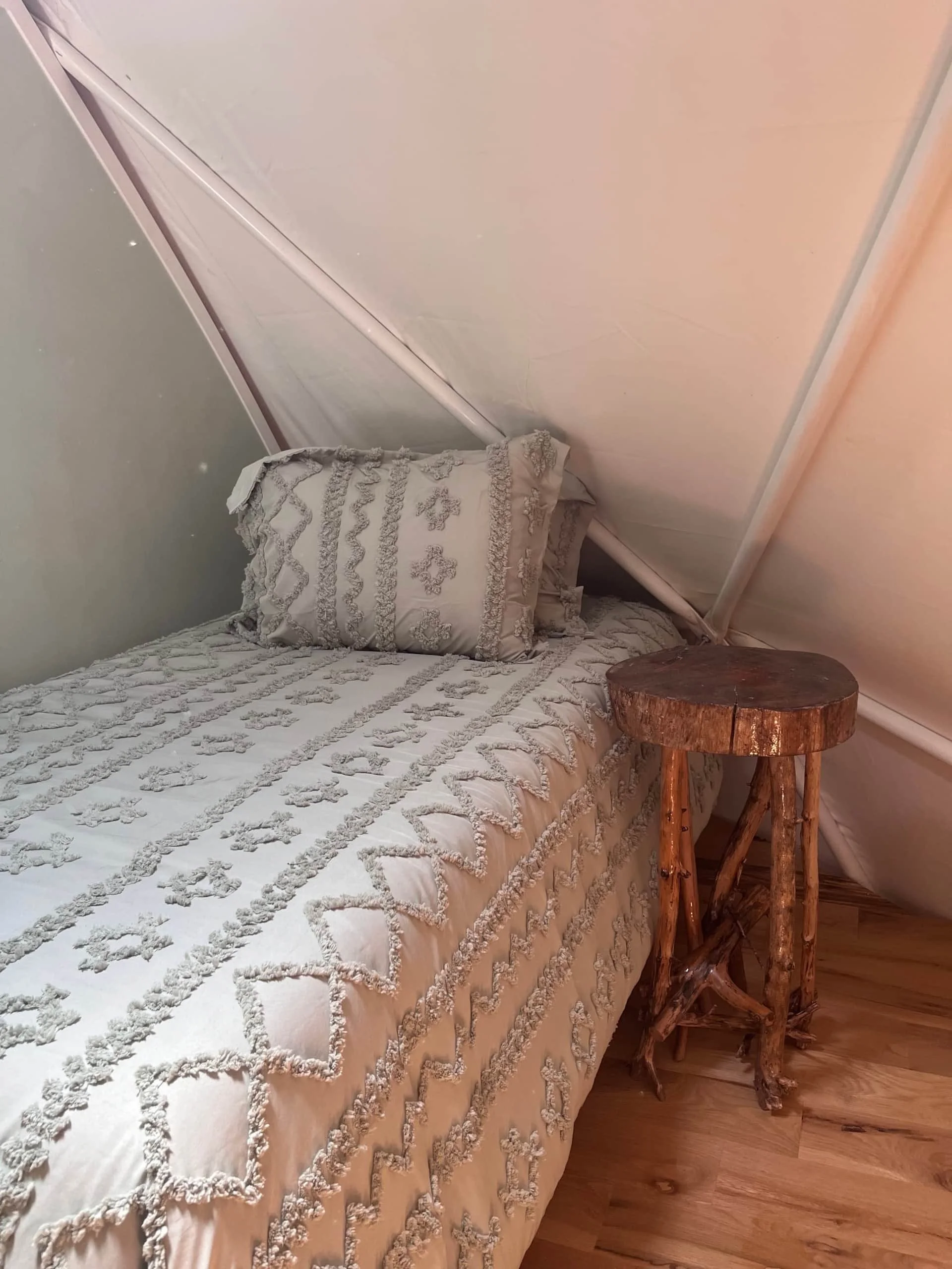 A bed with ornate white bedding and a matching pillow, a small rustic wooden side table, and a white wall with sloped ceiling.
