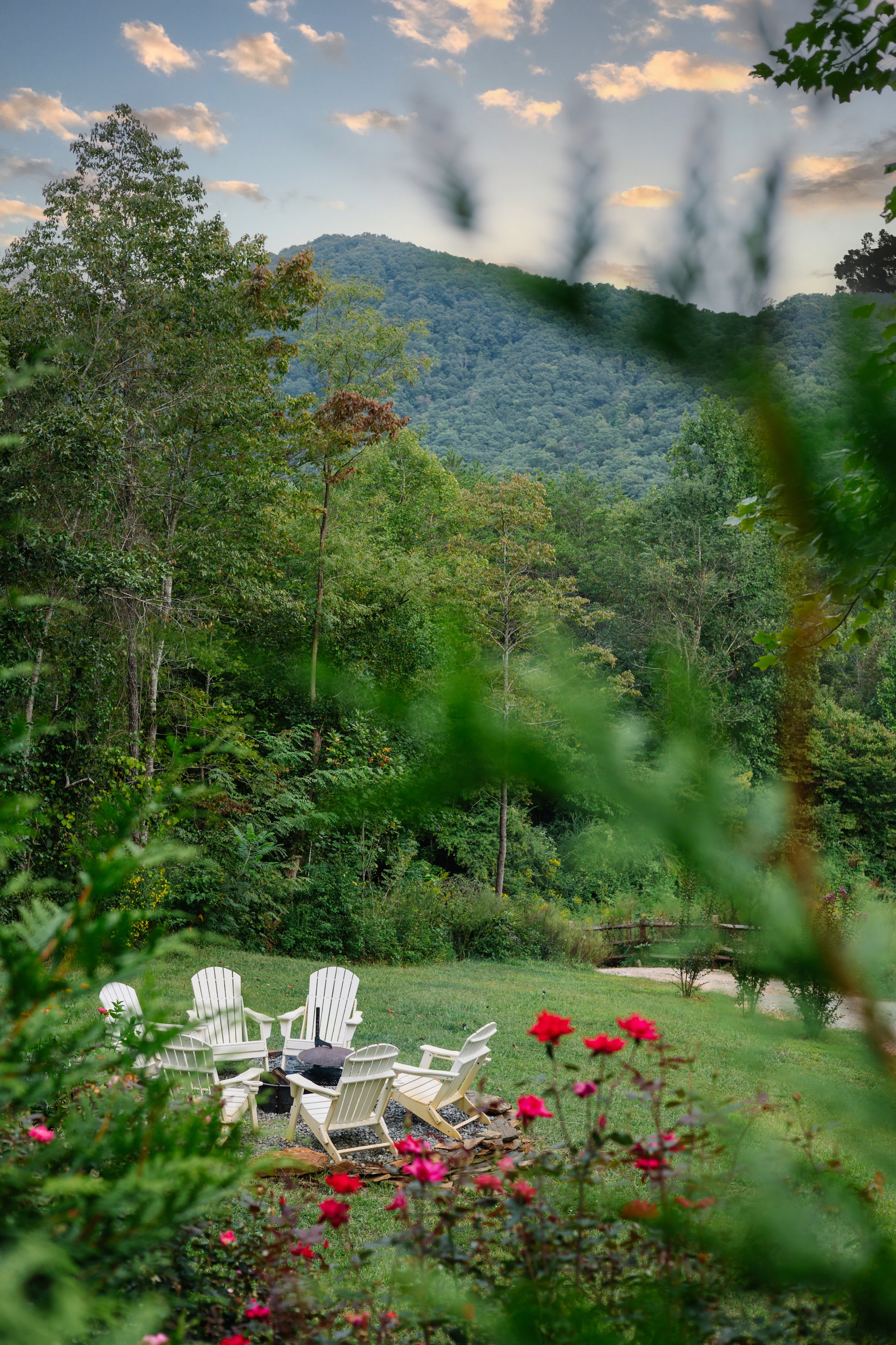 An outdoor scene with a circular arrangement of white Adirondack chairs on a grassy lawn, surrounded by vibrant pink flowers and lush green trees, with forested mountains in the background under a partly cloudy sky.