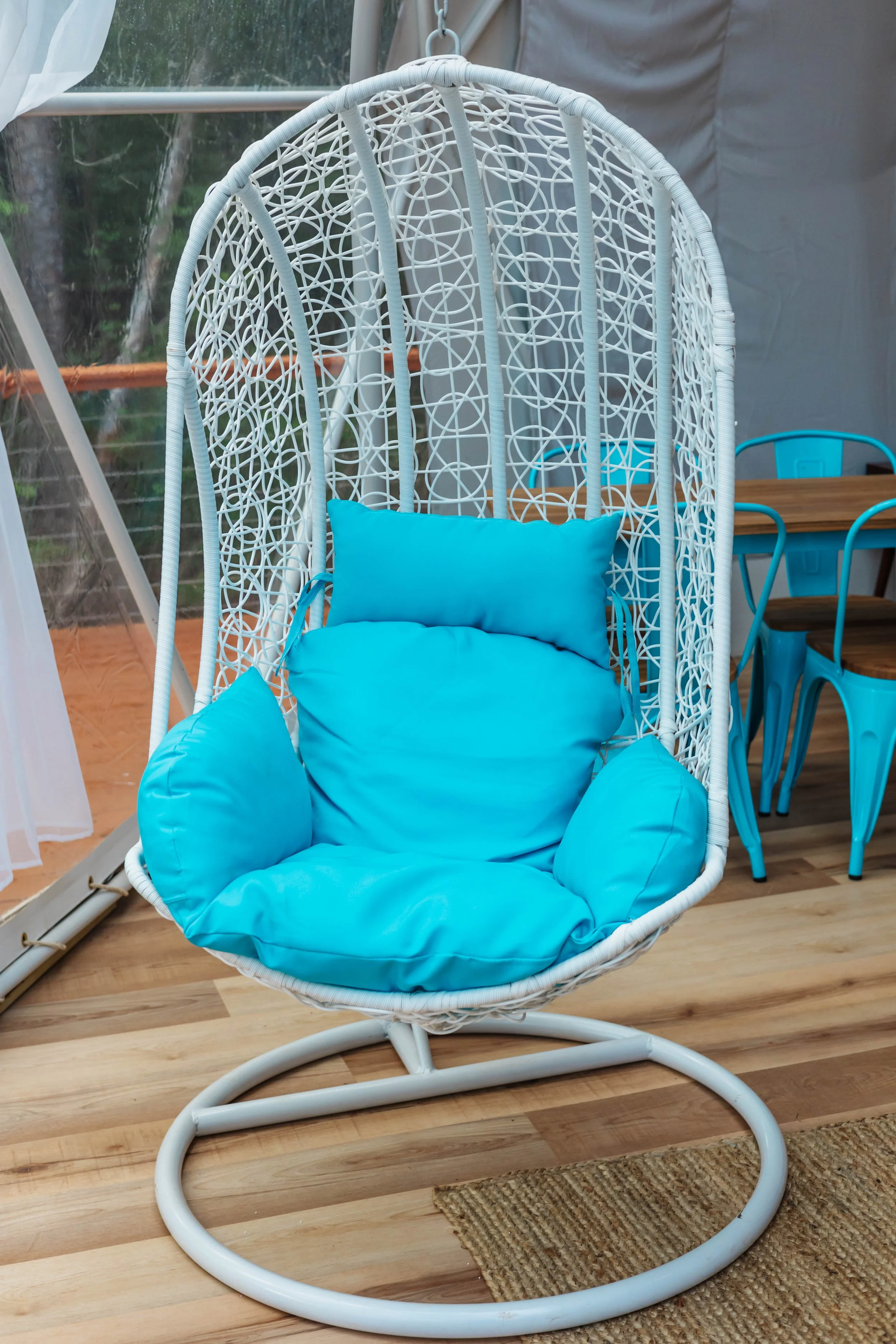 A white woven hanging chair with blue cushions inside, positioned on a wooden floor near a glass door. There are blue chairs and a wooden table in the background.