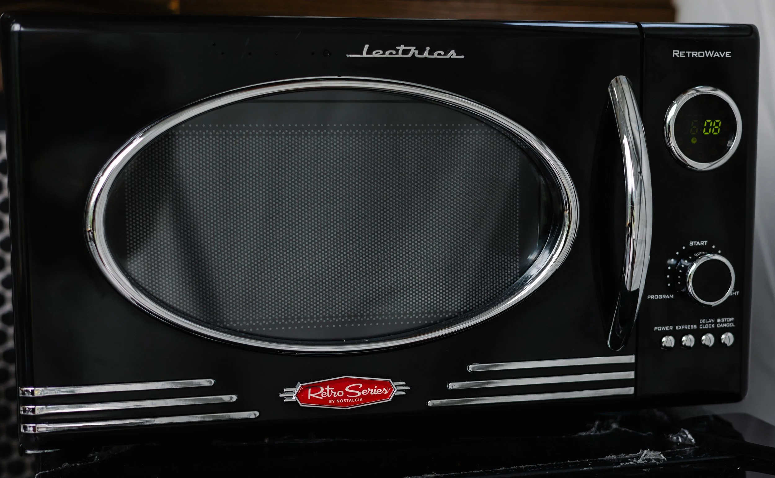 A black vintage-style microwave oven with silver accents, labeled 'Retro Series' by Nostalgia, featuring a digital clock display showing 6:08, a round control knob, and several small buttons on the right side.