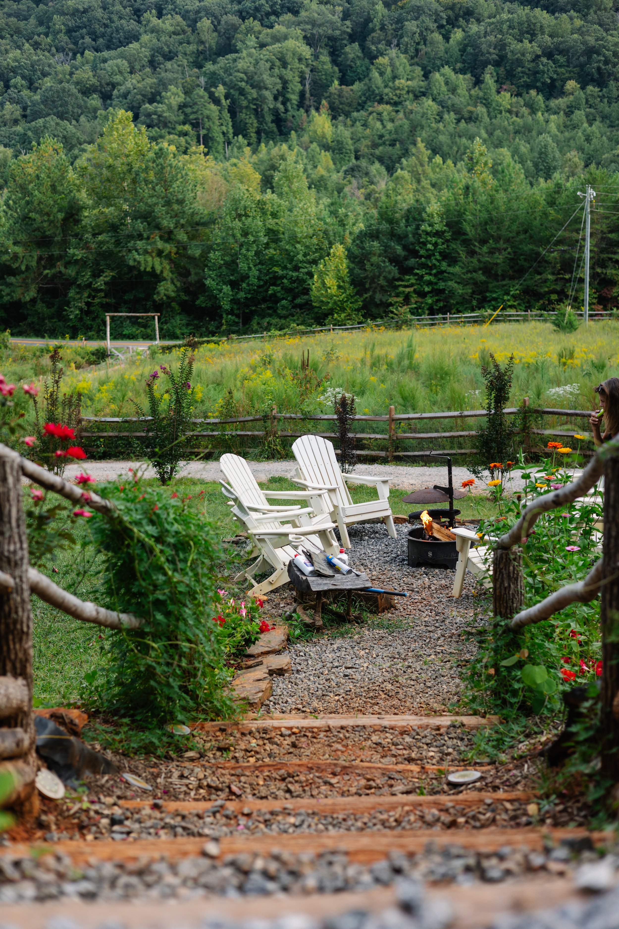 A cozy outdoor seating area with two white Adirondack chairs and a firepit, surrounded by flowers and a garden, with a mountain and lush green trees in the background.
