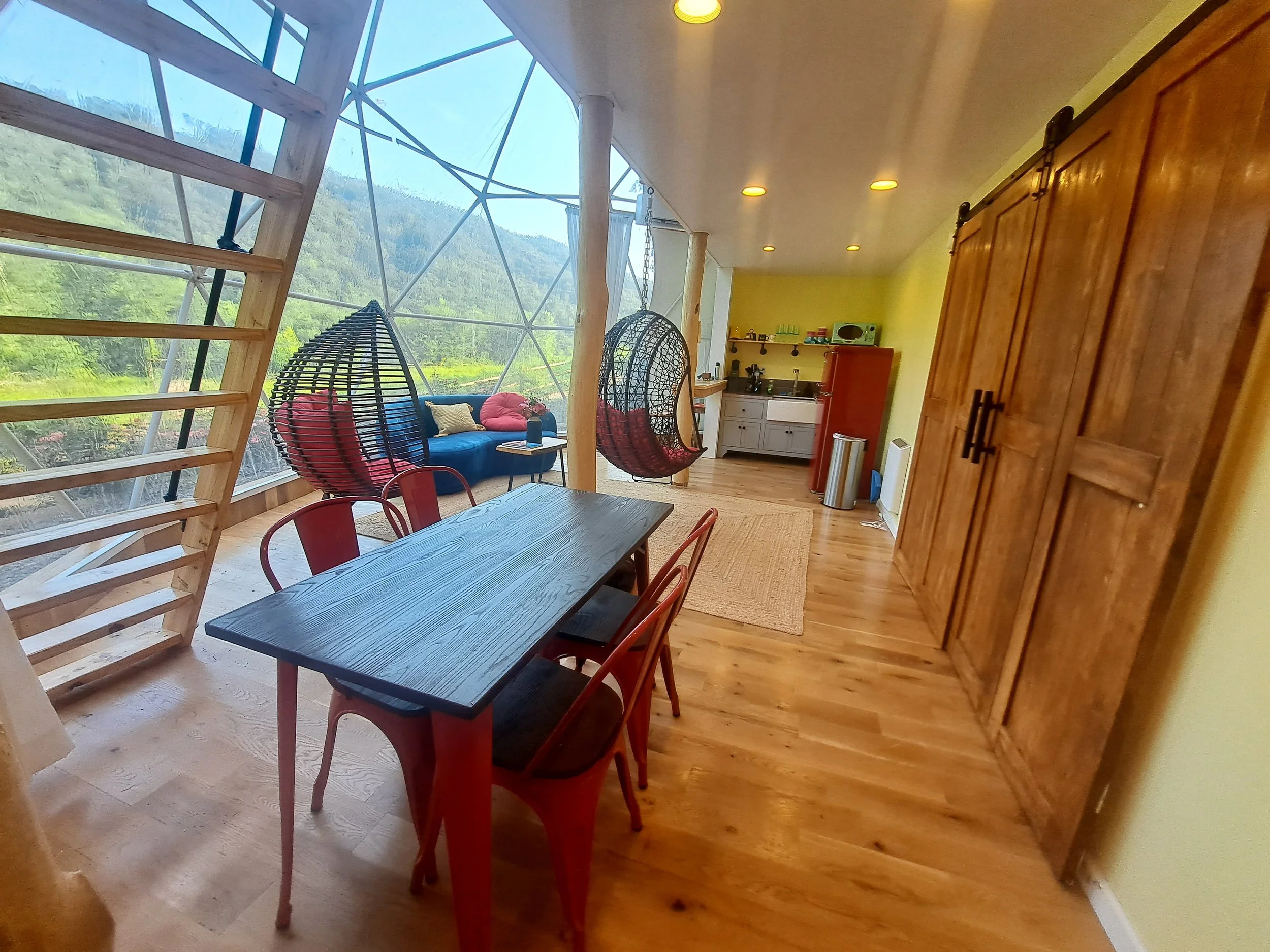 Interior view of a cozy living space with a large window showing outdoor greenery and mountains. The room has a wooden floor, a dining table with red chairs, hanging bubble chairs with red cushions, a small sofa with cushions, and a kitchenette in th
