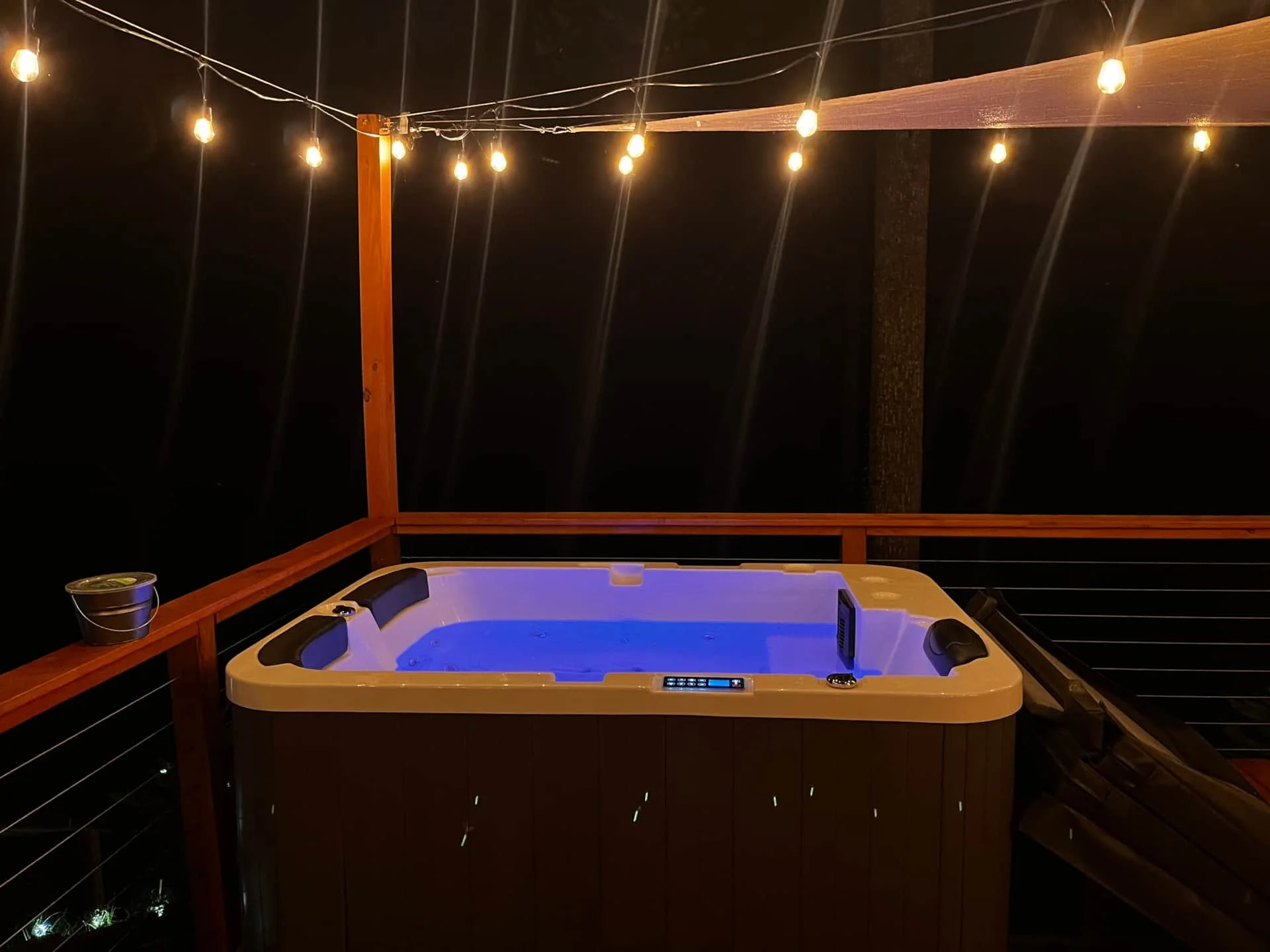 Outdoor hot tub illuminated with blue light, surrounded by wooden railing and string lights at night.