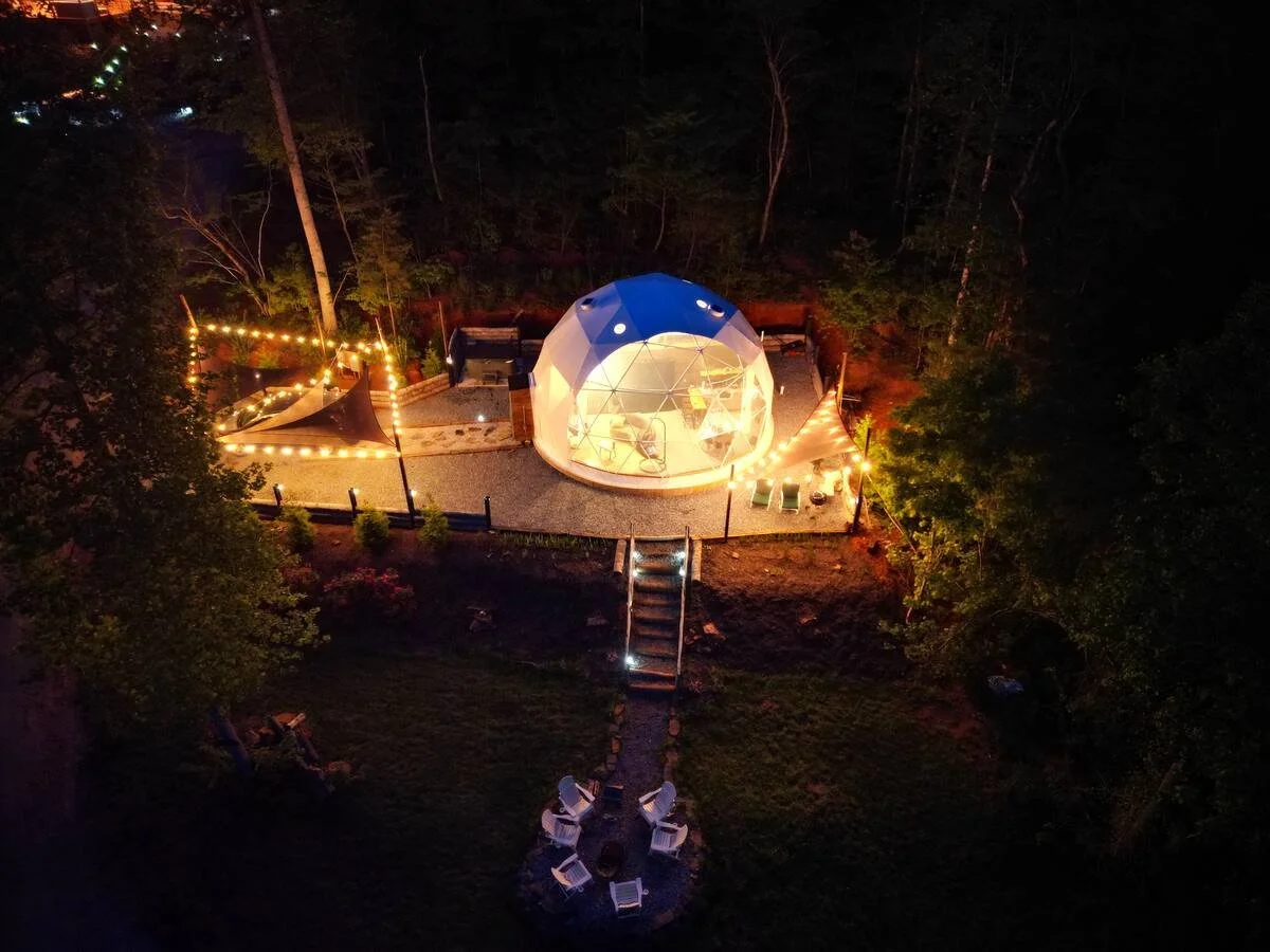 A transparent geodesic dome illuminated at night, situated in a wooded area. String lights create a cozy outdoor atmosphere with chairs and a path leading to the dome.