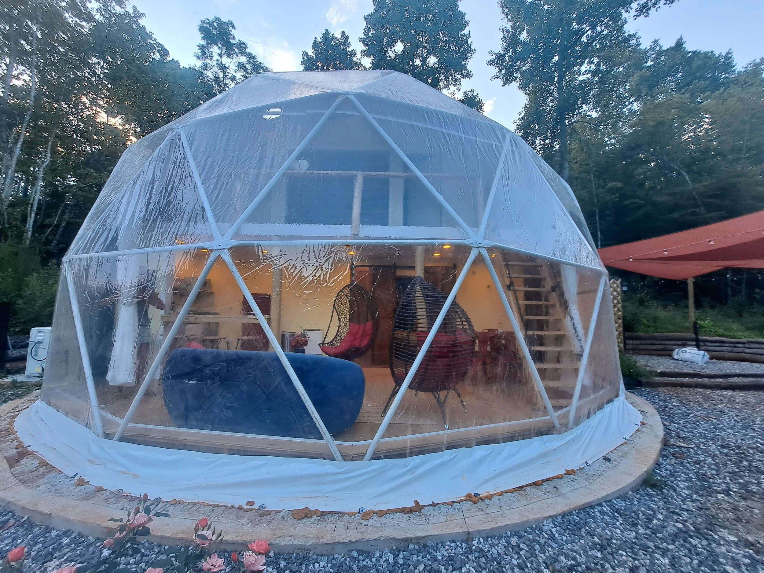 A transparent geodesic dome outdoor structure with visible interior furniture and seating, set in a natural wooded area with trees and a gravel ground.