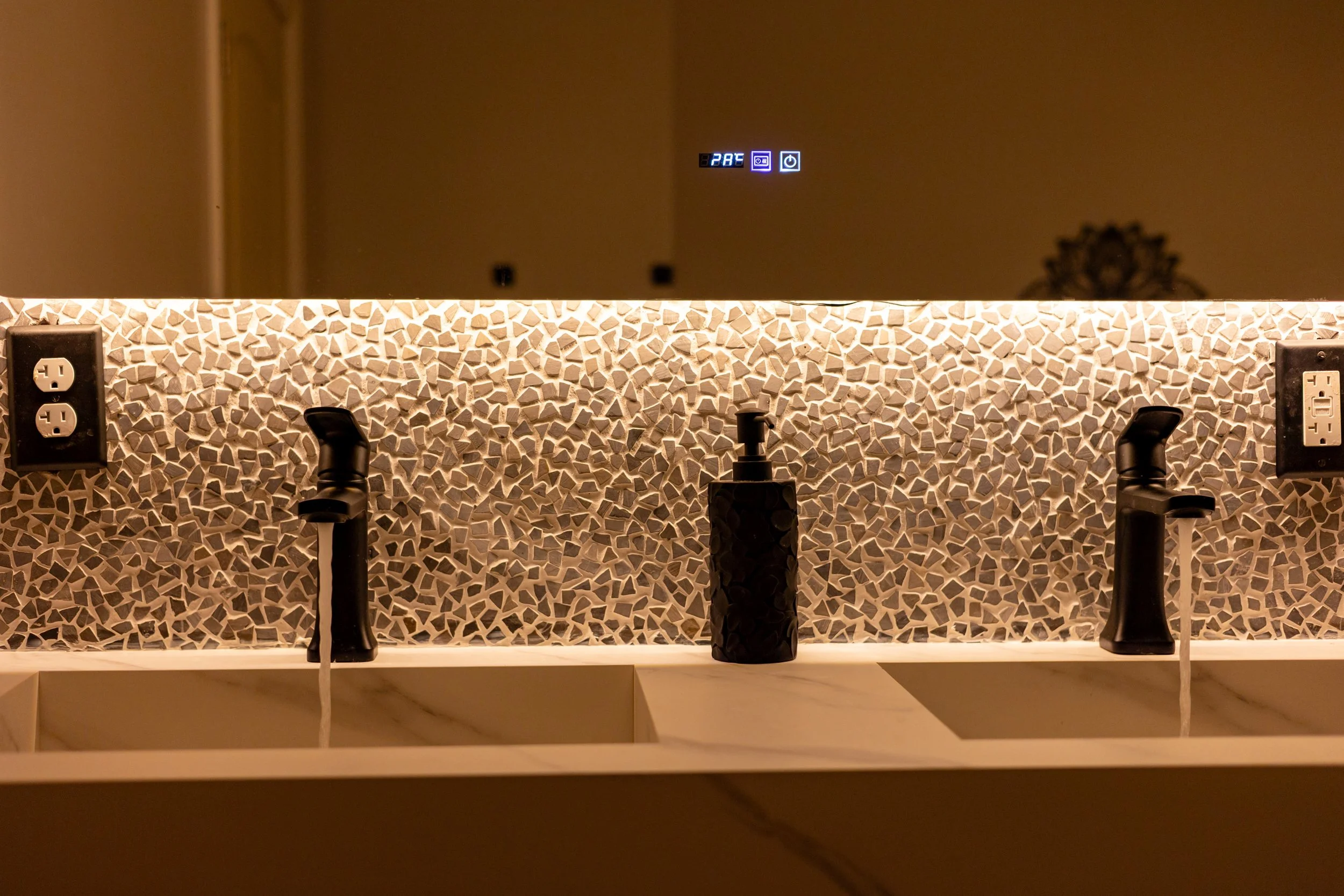 Bathroom sink with two black faucets, a soap dispenser, and a mosaic backsplash illuminated by warm lighting. There are electrical outlets on both sides and a mirror reflecting a digital thermostat on the wall.