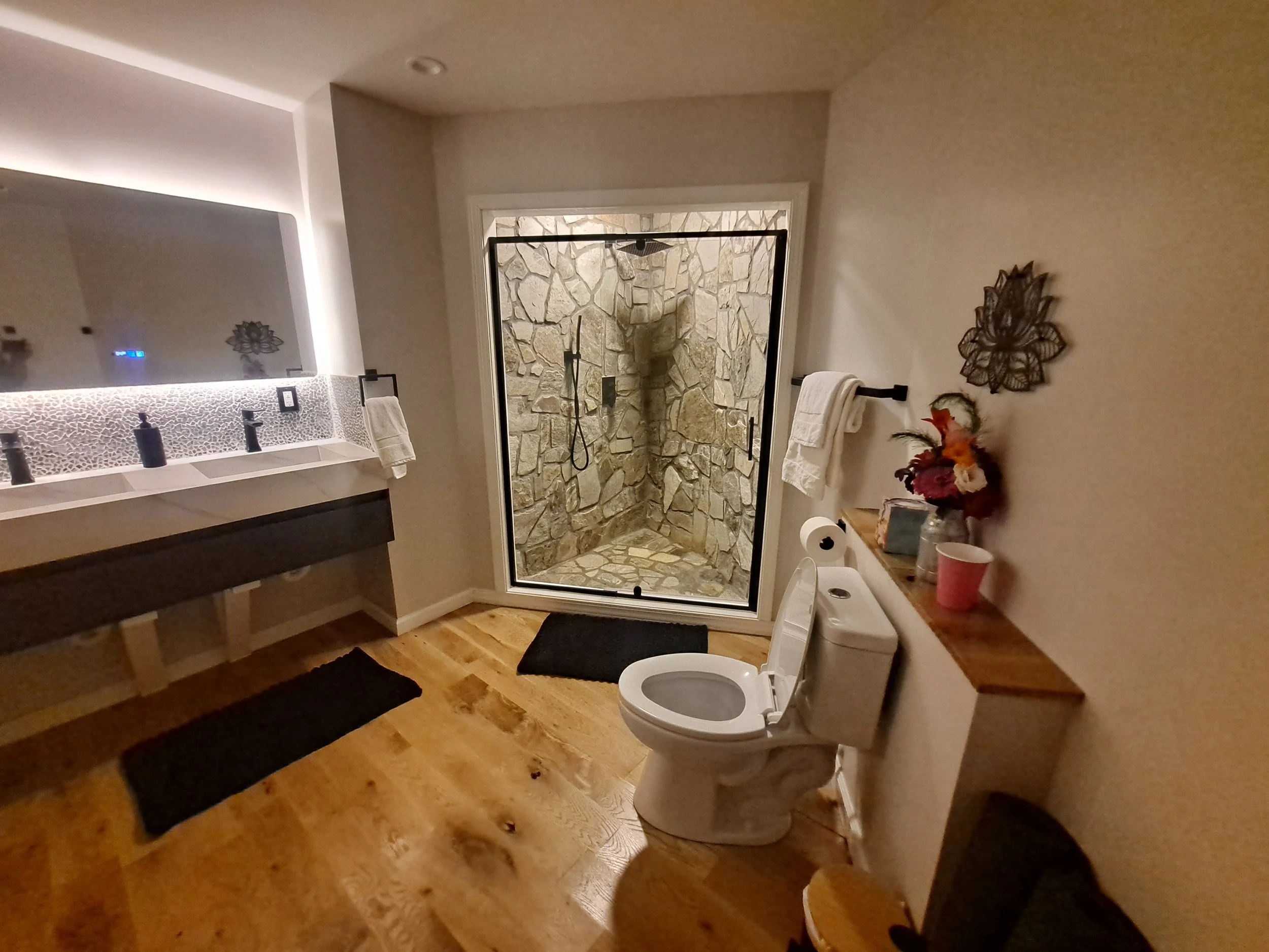 Bathroom with a glass shower enclosure with stone walls, a white toilet, and a wood vanity with a large mirror. Decor includes towels, a bouquet of flowers, and decorative wall art.