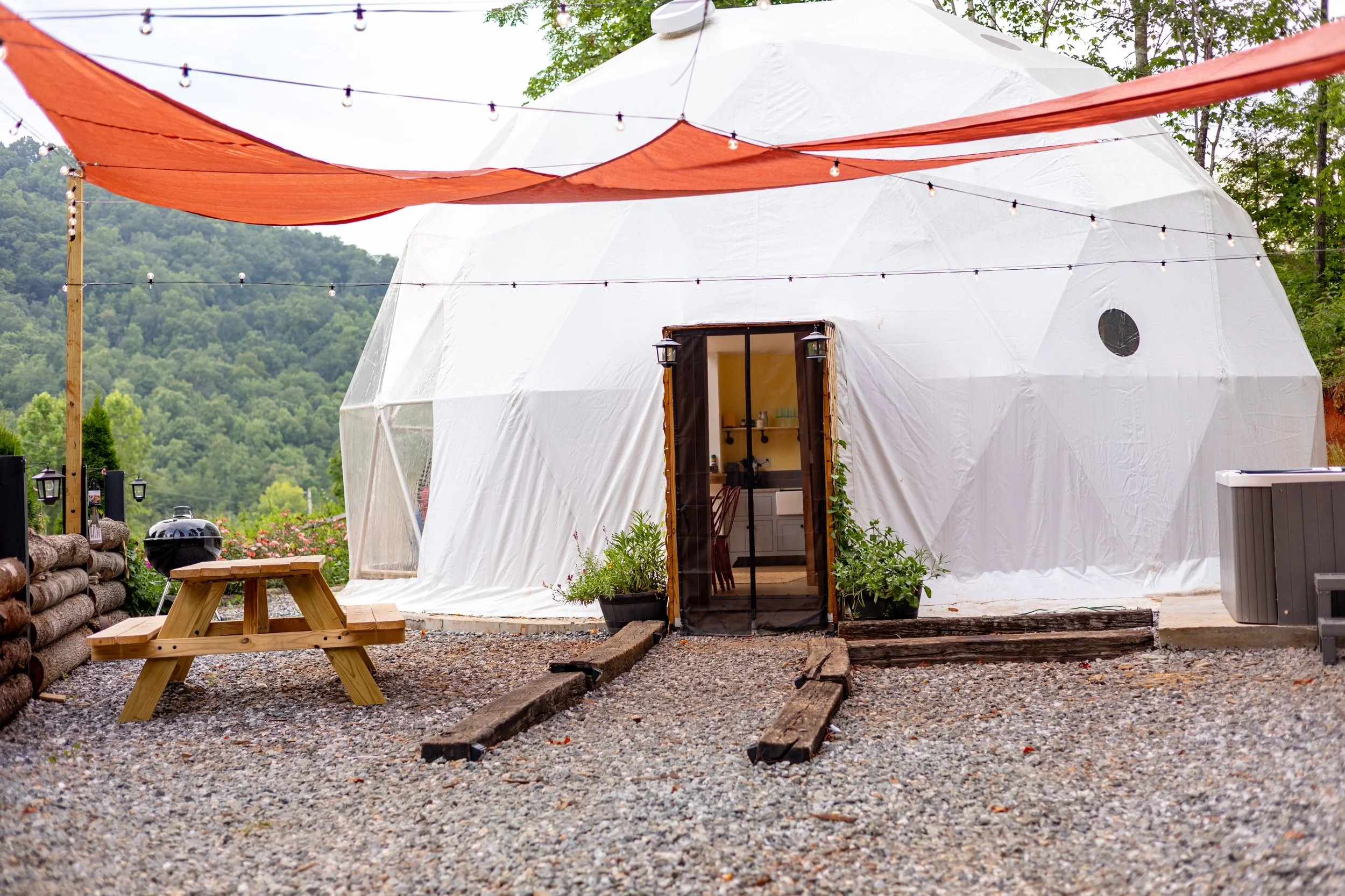 A geodesic dome tent with a wooden entrance door, surrounded by plants, outdoor string lights, a picnic table, a charcoal grill, and on a gravel surface, with green hills in the background.
