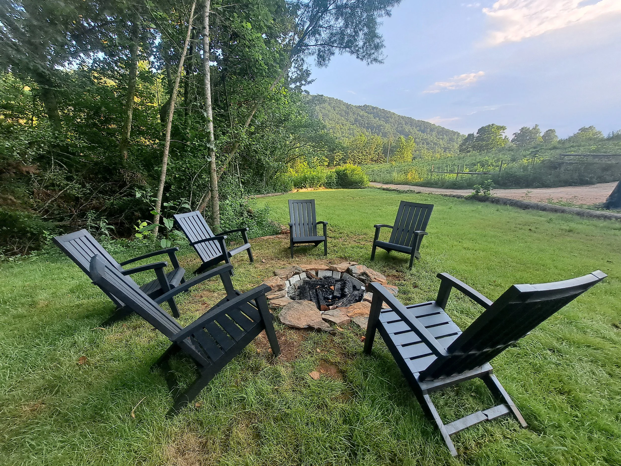 Six black Adirondack chairs arranged in a circle around a small stone fire pit in a grassy backyard with trees and a green hillside in the background.