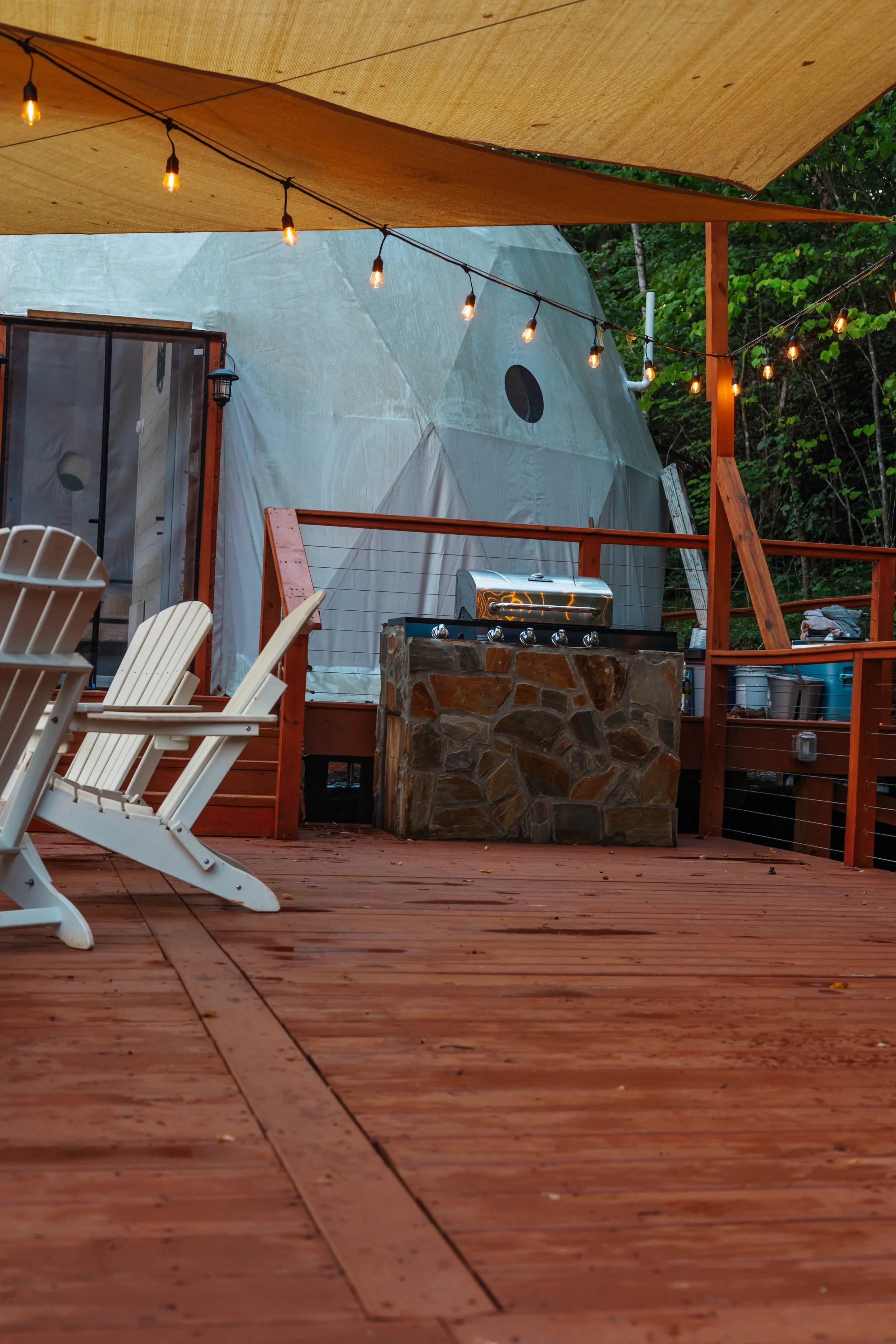 Outdoor deck with Adirondack chairs, a stone outdoor grill, string lights, and a large tent in a wooded backyard.