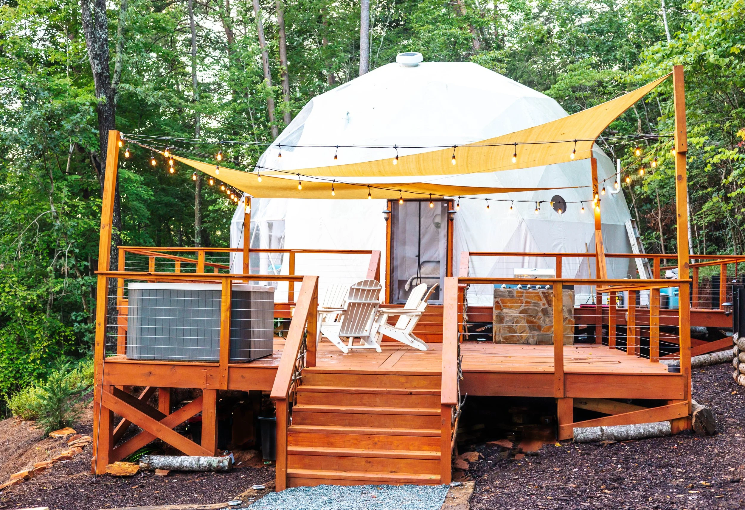 Wooden deck with Adirondack chairs and string lights, in front of a white geodesic dome surrounded by trees.
