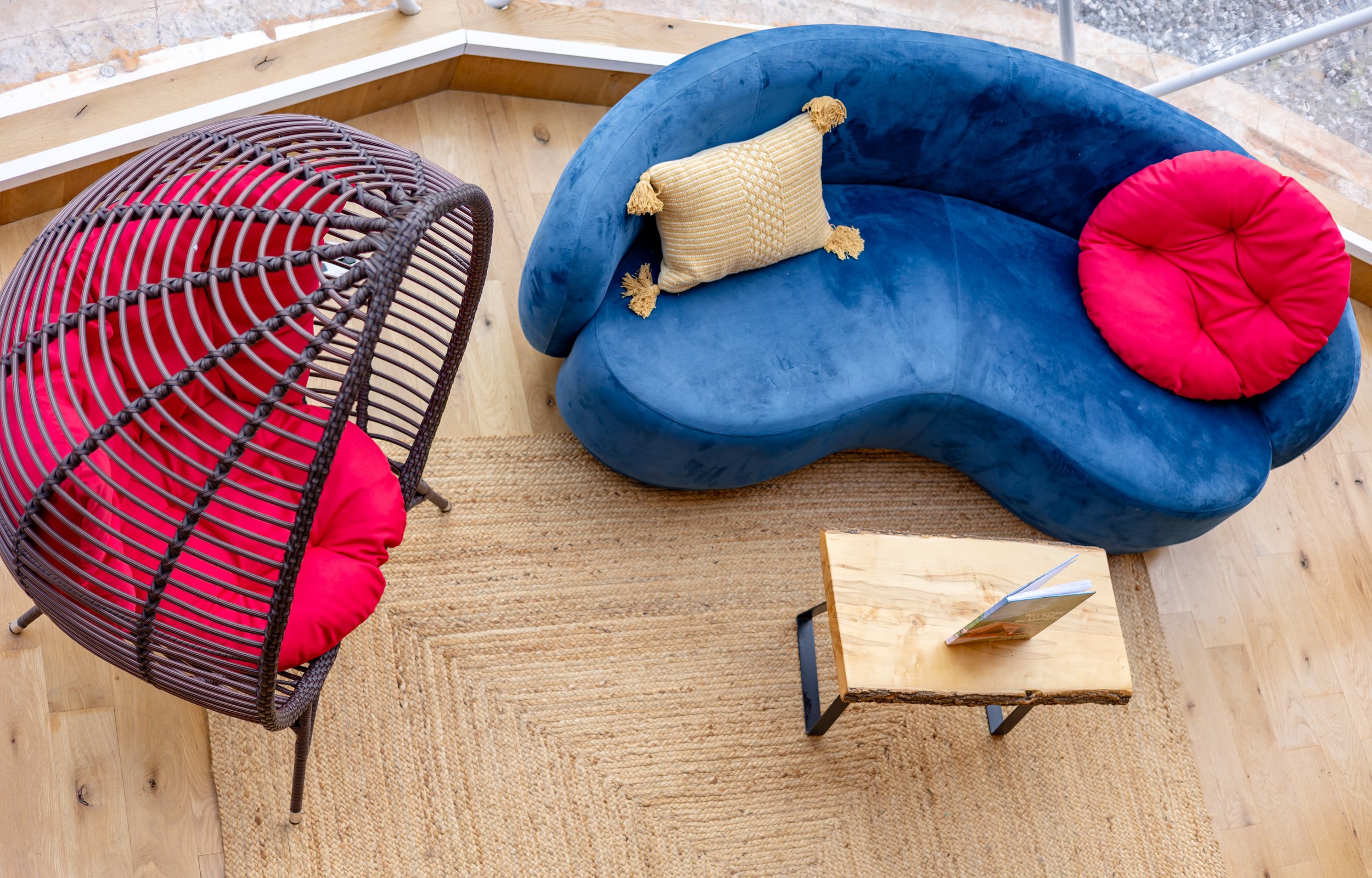 Indoor seating area with a curved blue velvet sofa, a wooden coffee table with a book, a wicker chair with a pink cushion, and a woven beige rug.