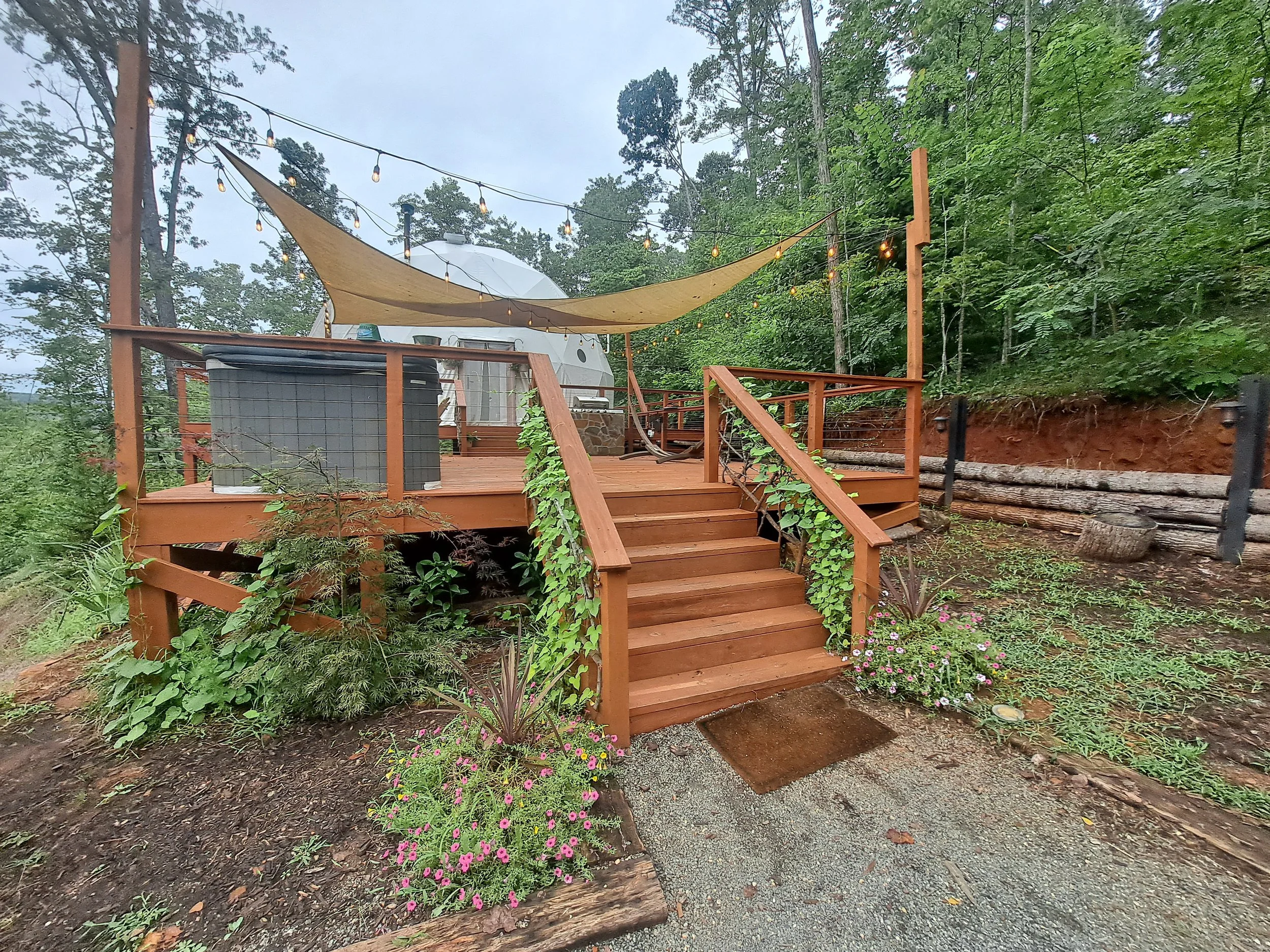 Wooden deck with stairs, string lights, and a sunshade, surrounded by greenery and flowers, with a small camper or trailer in the background.